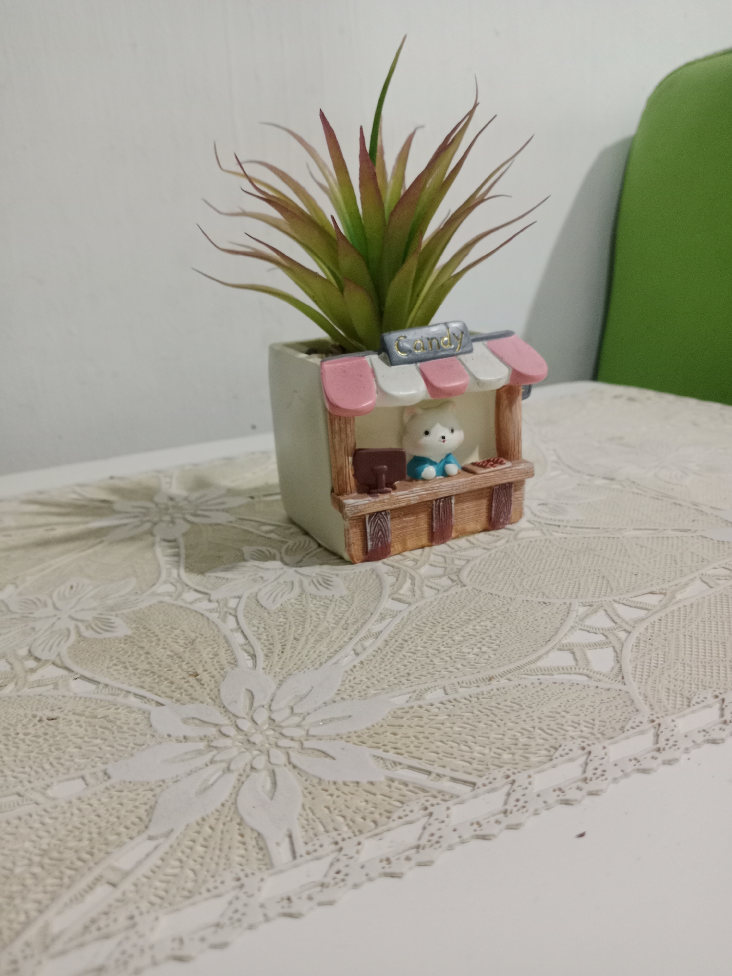 An artificial succulent plant with green and reddish-brown leaves is housed in a white pot that is designed to resemble a small candy shop. The shop has a pink and white striped awning, a wooden counter, and a miniature white cat figurine wearing a blue shirt behind the counter. The pot is placed on a white tablecloth with a detailed, floral lace pattern. The background consists of a plain white wall and a portion of a green chair. The image was taken indoors, likely on a table. The location in the city of Tambakcekur, Indonesia, is not ascertainable from the visual information provided in the image.