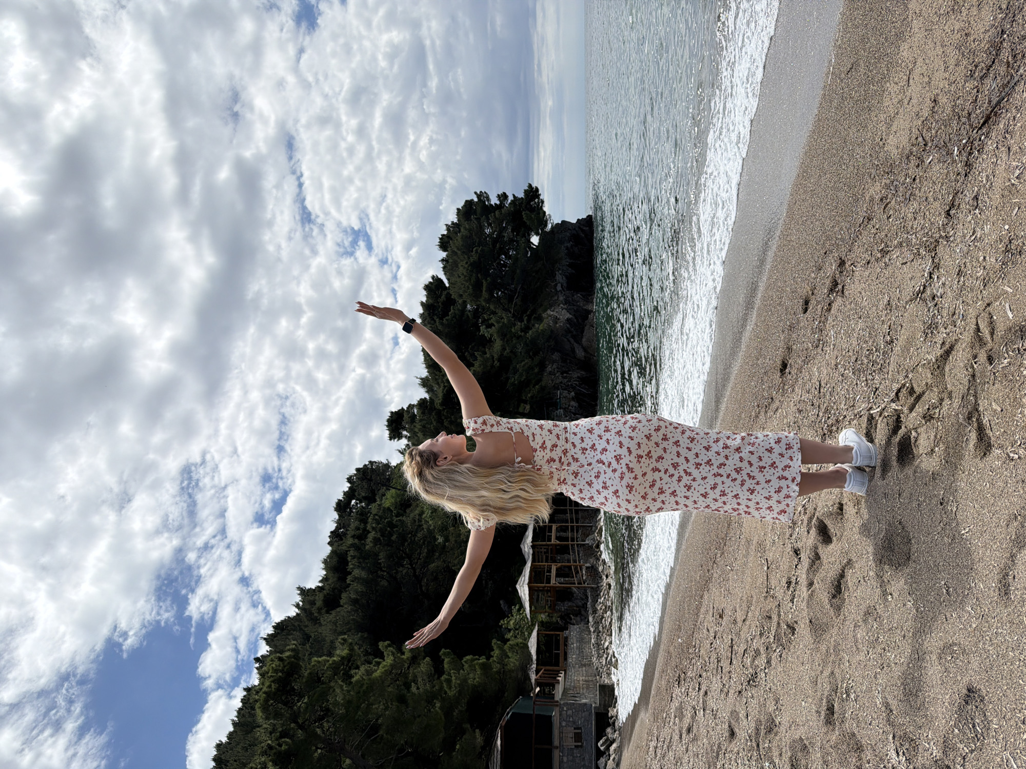A woman in a floral dress stands on the sandy shore of a beach with her arms raised. The sea stretches to the right side of the frame. Behind her, lush greenery covers the rocky terrain. A cloudy sky fills the upper part of the image. The scene is located in Petrovac centar, Montenegro.