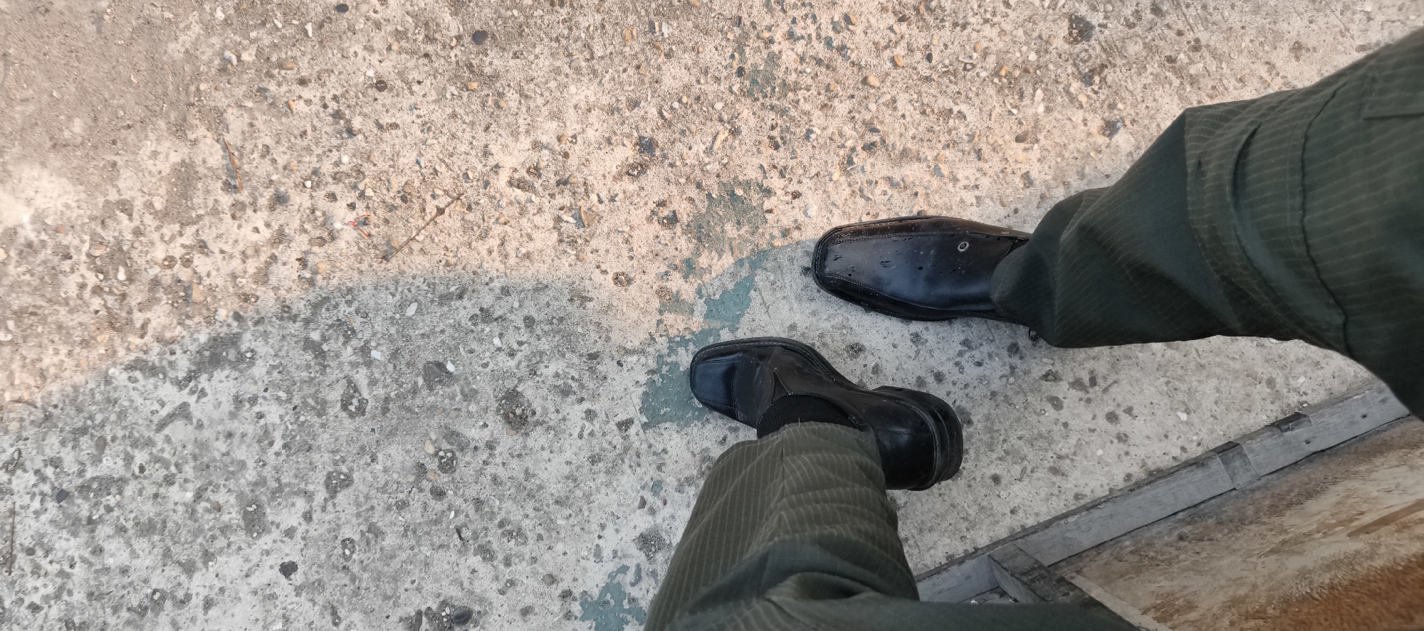 The image shows a low-angle, first-person perspective of a person's legs and feet wearing black, square-toed dress shoes and dark green corduroy pants. The person is standing on a concrete surface with visible pebbles and some teal-colored staining. A shadow of the person's legs is cast on the ground to their left. To the right, a wooden structure, possibly part of a bench or railing, is visible. The lighting suggests it is daytime, with sunlight illuminating the scene from the upper right. The image does not provide any specific landmarks or clues to pinpoint a location within Baru, Indonesia.