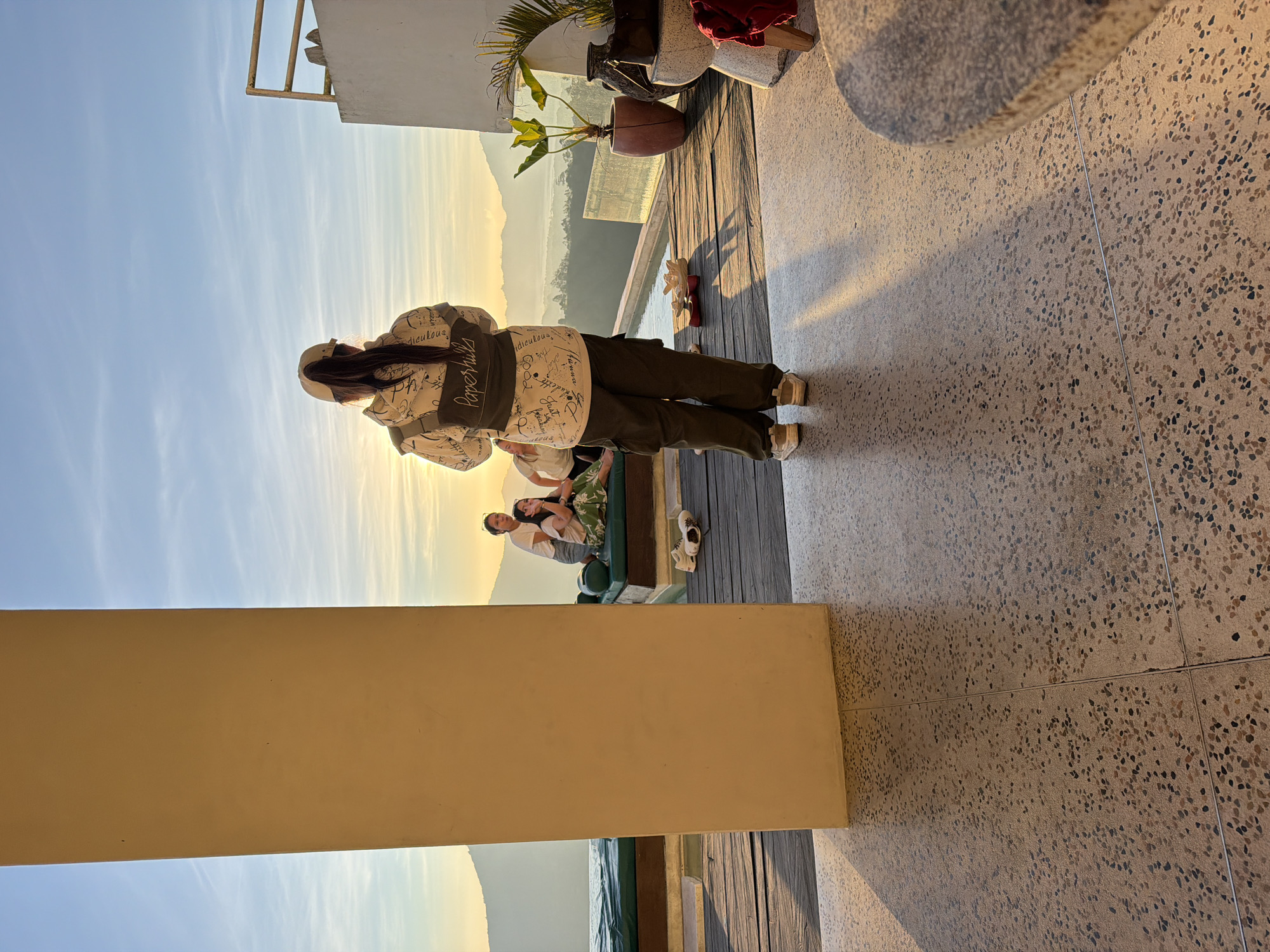 The image depicts a panoramic view from an outdoor terrace in Kintamani, Indonesia, likely at sunrise or sunset given the low-angle golden light. In the foreground, a person stands with their back to the camera, wearing a white hooded jacket with dark script, dark cargo pants, and a light-colored baseball cap covering a ponytail. They are positioned on a light brown terrazzo-style floor with dark speckles, which extends to the right where a rounded stone table edge is partially visible. A prominent beige rectangular pillar stands on the left, vertically bisecting the midground.
Beyond the pillar and to the left, three individuals are seated or lounging on dark green cushioned daybeds next to an infinity pool. One woman in a white t-shirt and patterned skirt looks left, while another in a white t-shirt looks right. Several pairs of shoes are scattered on the wooden deck adjacent to the pool.
The background reveals a vast mountainous landscape, characteristic of Kintamani's volcanic region, with a valley or lake partially obscured by atmospheric haze. The sky is clear with scattered light clouds, bathed in the warm, golden glow of the sun, which is visible as a bright flare behind the main subject. To the far right, near the pool's edge, a large reddish-brown potted plant and a dark ceramic vase are visible next to a white concrete structure featuring a yellow metal railing. Long shadows are cast across the foreground floor by the early/late day sun.