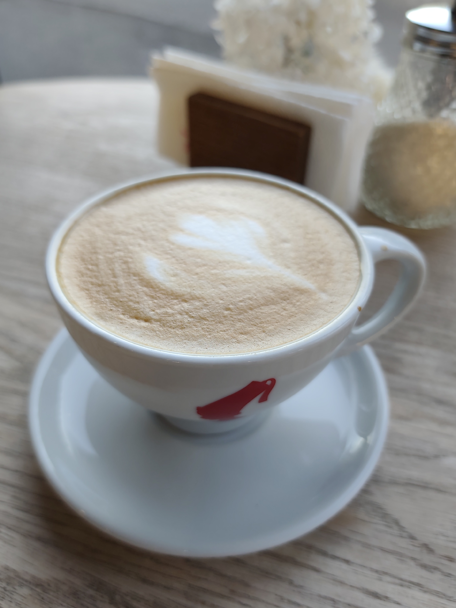 A close-up, eye-level shot focuses on a white ceramic cup filled with a frothy cappuccino, featuring latte art in the shape of a heart. The cup rests on a matching saucer, both placed on a light-colored wooden table. In the background, slightly out of focus, is a napkin holder with white napkins and a brown dispenser, along with a glass sugar shaker containing white sugar. The scene is indoors, likely in a cafe or restaurant, given the presence of coffee service items. The overall lighting is soft, suggesting ambient interior lighting. The specific location in Sochi, Russia, cannot be determined from the visual information provided. There are no people visible in the image.
