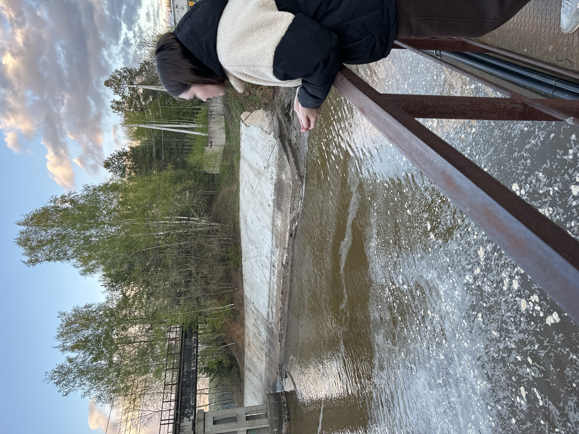 A woman stands on a rusty metal bridge overlooking a body of water, likely a river or canal, in Chaikovsky, Russia. The water is turbulent with white foam. The woman is wearing a jacket with a black and white color block design. She is looking down at the water and appears to be interacting with it using her hand. A concrete embankment lines the water's edge on the opposite side of the bridge. Trees and power lines are visible in the background. The sky is cloudy.