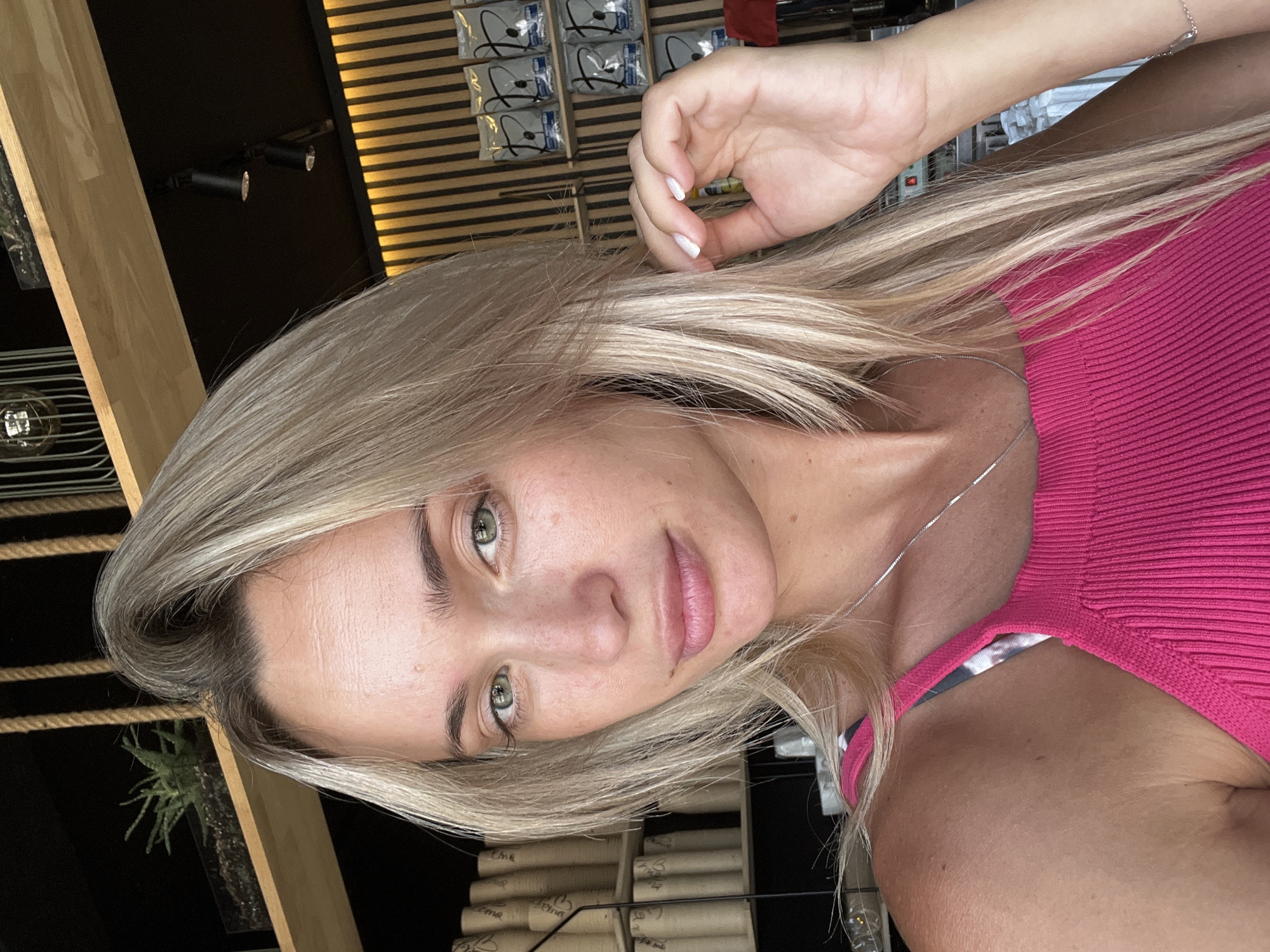 A woman with blonde hair and green eyes is pictured in a close-up shot, looking directly at the camera. She is wearing a bright pink ribbed tank top and a silver necklace. Her left hand is touching her hair, and her right hand is partially visible on the right side of the frame. The background appears to be indoors, with shelves filled with cylindrical objects and some greenery visible on a higher shelf. The lighting suggests it is daytime. The image is taken from the chest up, focusing on the woman's face and upper body.