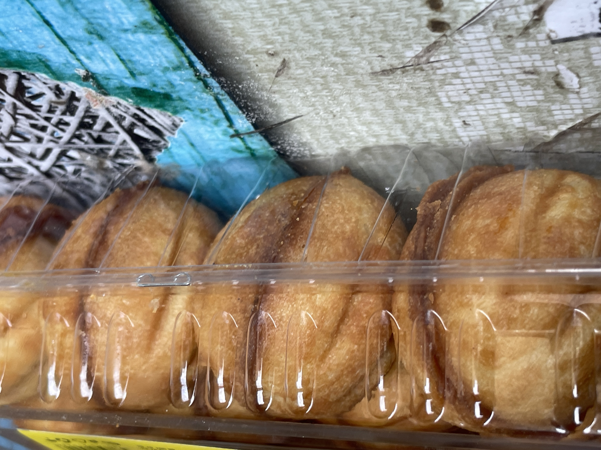 A close-up, eye-level, horizontal shot shows a clear plastic container with a yellow label at the top, filled with rows of golden-brown, baked goods. The baked goods appear to be pastries or bread rolls, with a textured, slightly flaky surface. They are arranged in two stacked layers, with the front row clearly visible and the back row partially obscured. The plastic container has indentations that create divisions between the individual items. The background beneath the container is a patterned surface with a light beige and white checkered design transitioning into a vibrant turquoise and grey textured pattern. The lighting is bright, casting reflections on the plastic. There are no people or any identifying landmarks visible in the image. This image was captured in Tashkent, Uzbekistan.