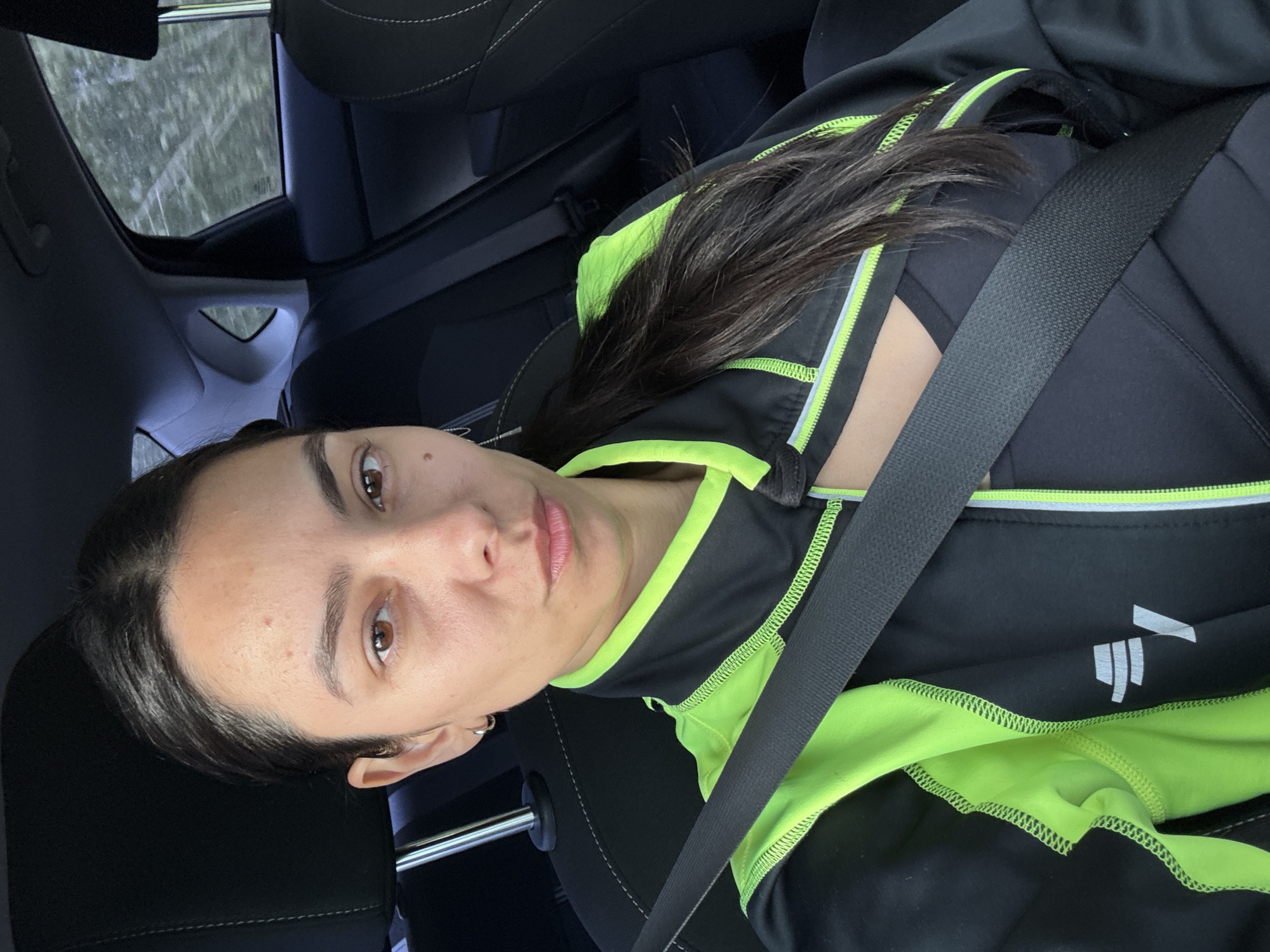 A woman is in a car, wearing a black jacket with neon green trim and a seatbelt. The car appears to be in motion, as blurry trees are visible through the window. The photo does not contain information regarding the location in the city of Chaikovsky, Russia.