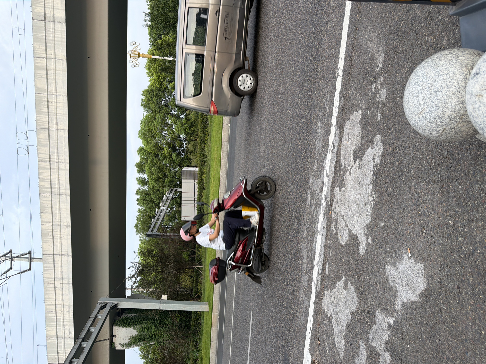 The image depicts a street scene in the city of Jiangjin, China. In the foreground, the asphalt road surface features a white painted line and several irregular patches of lighter colored material, possibly oil stains or wet spots. A discarded cigarette butt is visible on the road. In the middle ground, a person wearing a pink helmet and a white t-shirt is riding a red electric scooter. The scooter has a dark red storage box mounted on the back. To the right, a grey minivan is traveling in the same direction. The background is dominated by lush green trees and a large elevated concrete structure, likely a highway overpass, with electrical wires running overhead. The sky is overcast.