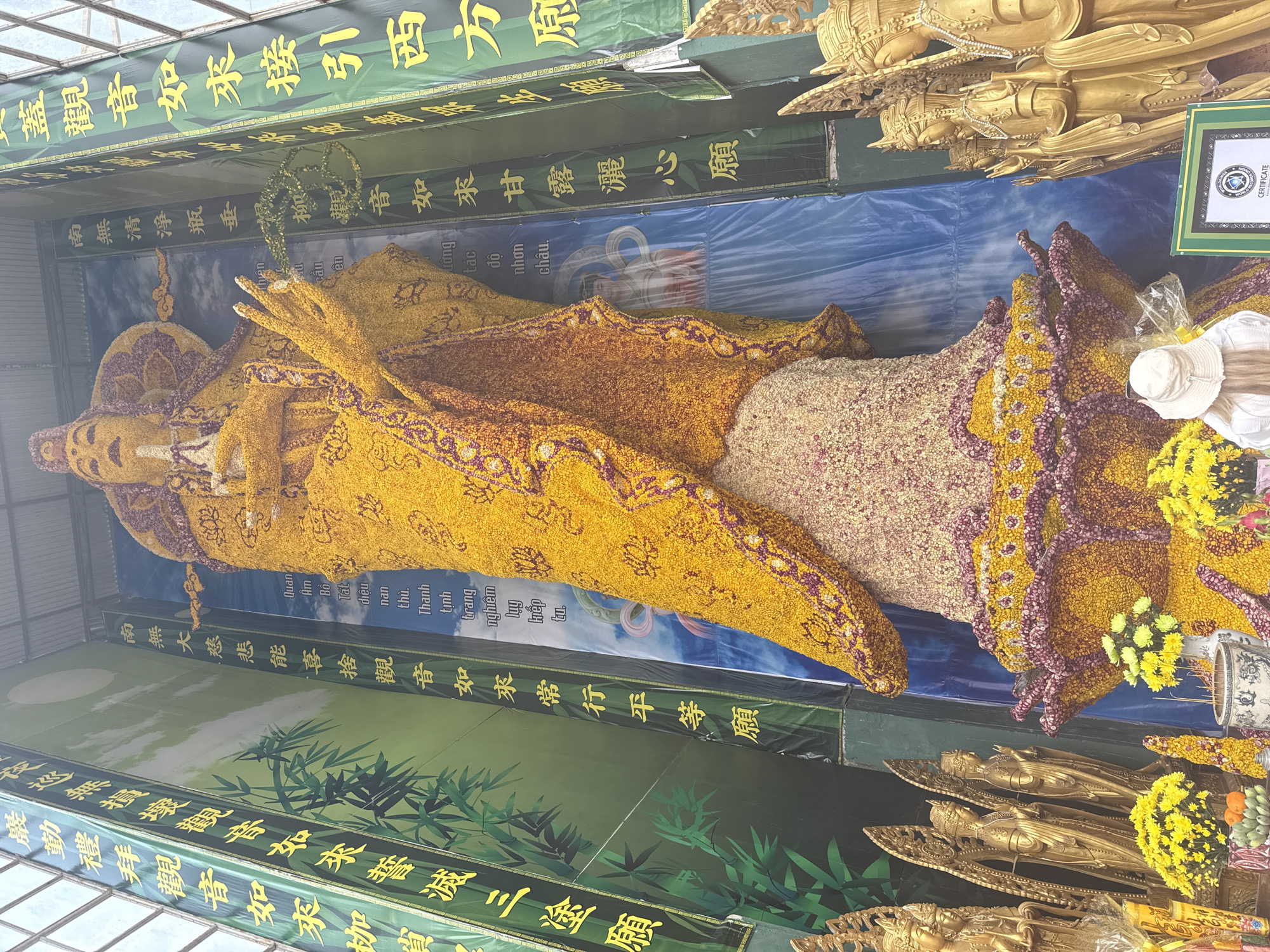 The image shows a large reclining Buddha statue adorned with yellow, purple, and white flowers. The statue lies horizontally, with the head to the left. The face and hands are covered in yellow flowers, while the robe has alternating bands of yellow and a lighter, possibly white or pink-tinged, floral covering. A garland of purple and yellow flowers defines the edge of the robe. The statue is displayed indoors, under a translucent roof. Green banners with yellow Chinese characters line the top and bottom of the display. Behind the statue is a blue banner with an image of water and white text. Golden Buddha statues are located below and to the right of the main statue. An official certificate is visible in the top right corner. The statue is in Xuân Thành, Vietnam.