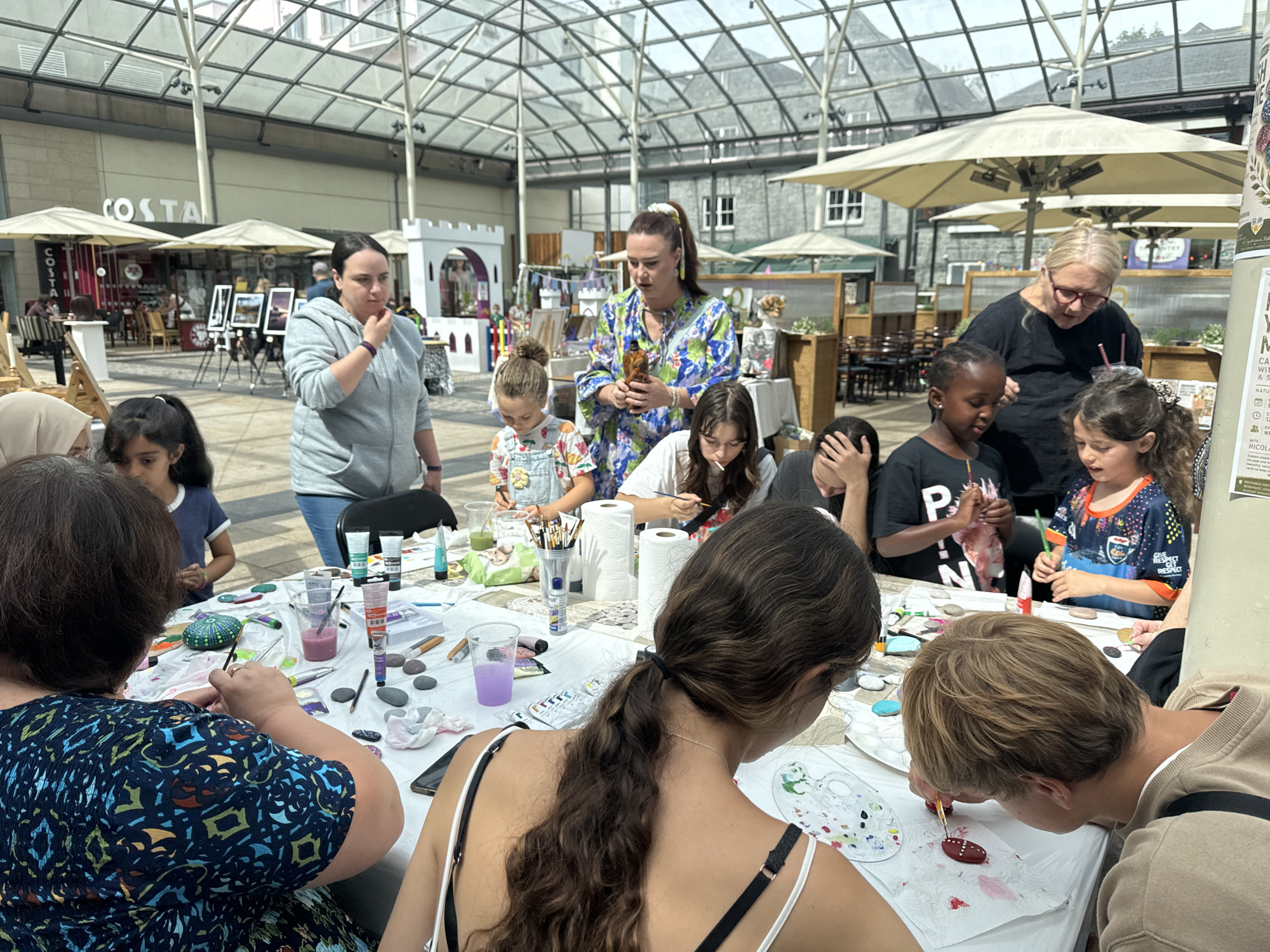 The image captures a vibrant arts and crafts workshop taking place in a covered outdoor area within the Kilkenny Design Centre in Kilkenny, Ireland. The scene is bustling with activity as a group of people, predominantly children and a few adults, are engaged in painting stones.
In the foreground, a woman with long, dark, wavy hair tied back is focused on painting a stone, her back to the camera. To her right, a young boy with blonde hair leans over a stone, also painting. Other children are visible around the table, some actively painting, others observing. One young girl with dark hair and a blue and orange t-shirt is holding a paintbrush and looking at her artwork. A woman with blonde hair and glasses, likely an instructor, is standing and appears to be guiding a young girl with dark skin. Another woman in a gray hooded sweatshirt stands in the midground, looking towards the activity. A woman in a colorful floral dress holds a bottle and appears to be demonstrating something.
The table is covered with a white cloth and is laden with art supplies including various tubes of paint, brushes in a container, water cups, paper towels, and numerous stones, some already decorated. The overall atmosphere suggests a creative and engaging event.
The background reveals the architecture of the Kilkenny Design Centre, with a glass-paneled roof allowing natural light to illuminate the space. Umbrellas are visible, suggesting an outdoor or semi-outdoor setting. In the distance, glimpses of shops and seating areas, including a Costa Coffee shop, can be seen, indicating the location is within a commercial and cultural hub. The event is taking place during daytime, under bright conditions.