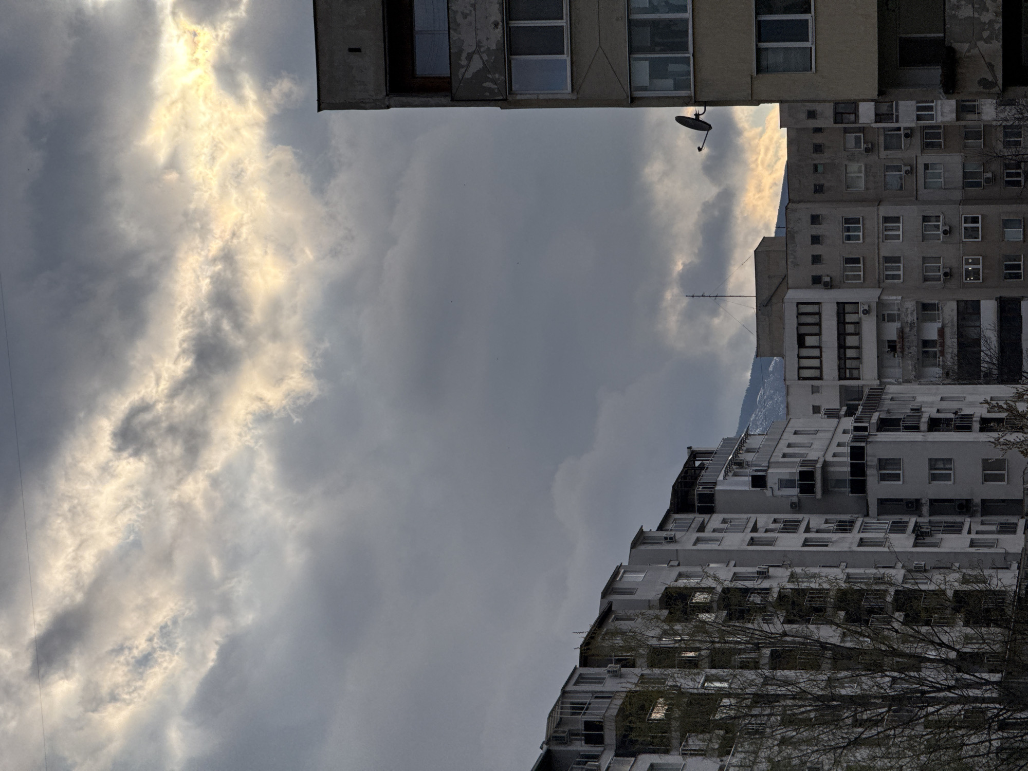 The image shows a view between buildings in Sofia, Bulgaria, looking up at a cloudy sky. The buildings are Soviet-era apartment blocks, with visible wear and tear. There is a satellite dish on the roof of one building. The sky is overcast with some brighter patches, suggesting the sun is behind the clouds. Branches of a tree are visible at the bottom right of the frame.