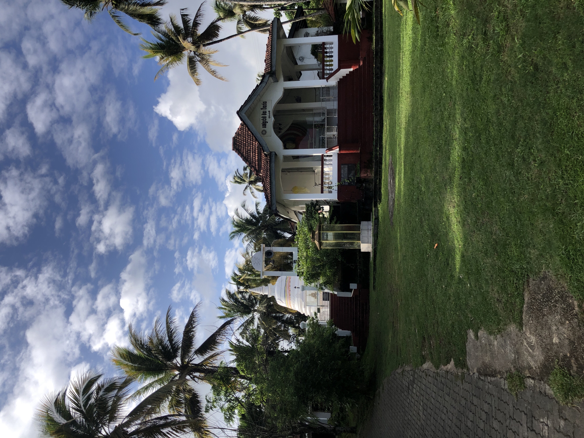 This image depicts a religious complex in Karandugoda, Sri Lanka, under a bright blue sky partially covered by scattered white cumulus clouds. Numerous tall palm trees with green fronds are prominent throughout the scene, framing the structures on the left, right, and in the background.
The foreground is dominated by a lush green lawn. Along the lower left, a paved pathway constructed from interlocking gray blocks curves towards the upper left, adjacent to a strip of dark earth.
Two main architectural structures are visible in the mid-ground. On the left, a white stupa (dagoba) with a golden finial rises from a raised platform, accessed by red-painted stairs featuring white railings. To the right of the stupa, a white structure incorporating a large bell is positioned. Further right and slightly forward of the stupa, a tall, slender, golden-colored display case rests on a white square base on the lawn.
On the right side of the mid-ground, a white building with a distinctive red tiled, multi-gabled roof is situated. This structure, consistent with a Buddhist shrine or temple, features a prominent front entrance with multiple red-painted steps and white railings leading to an open veranda supported by white columns. Sinhala script is visible above the entrance. A large, partially visible figure, likely a Buddha image, is discernible inside the veranda behind a protective barrier. Various green bushes and smaller trees are interspersed around the buildings and across the grounds. No individuals are present in the depicted scene.