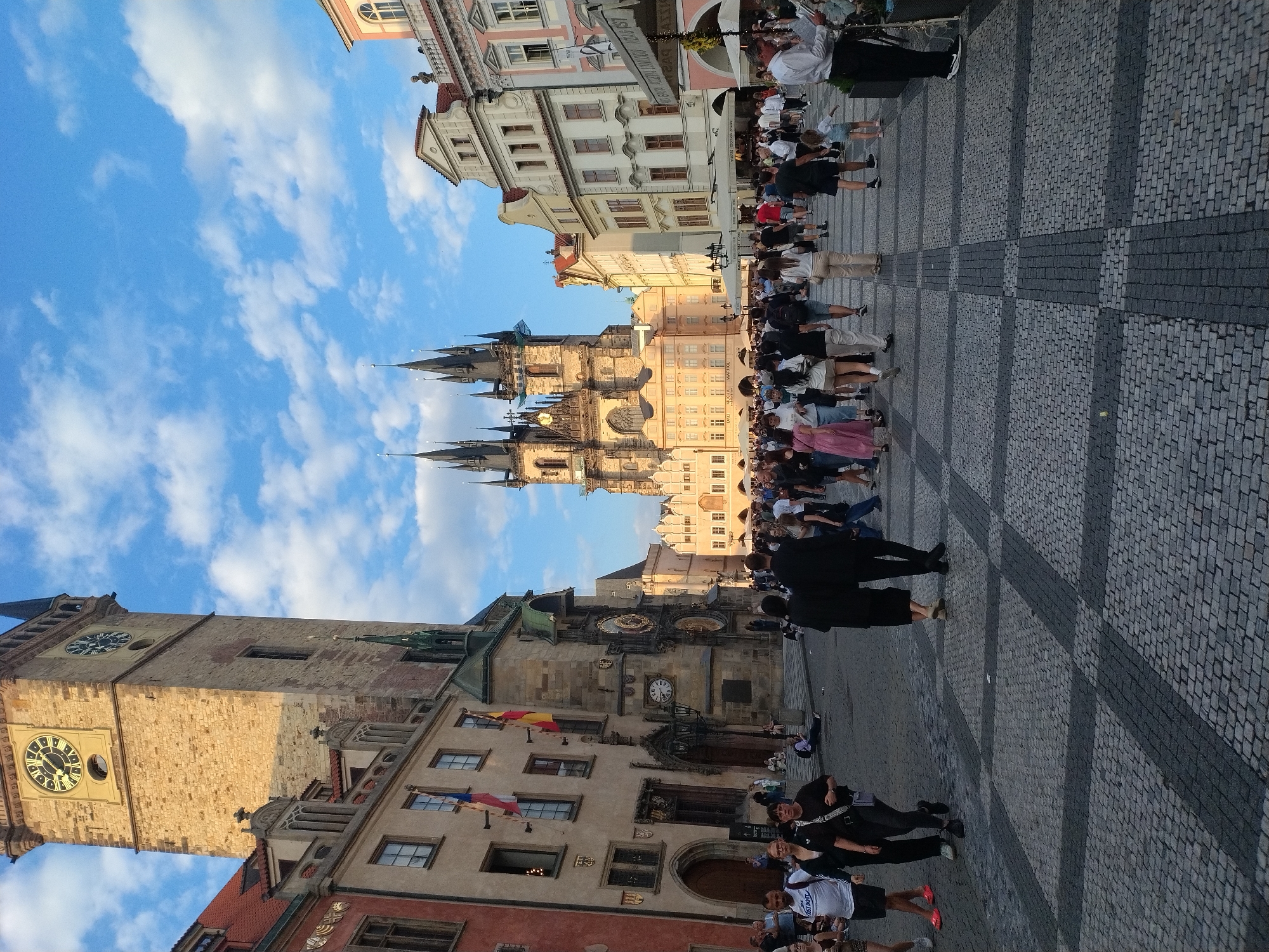 The image captures a view of Prague's Old Town Square, Czechia, with the Old Town Hall Tower and the Church of Our Lady before Týn prominently featured. The sky is blue with scattered white clouds, suggesting fair weather. A cobblestone square forms the foreground, with a checkered pattern of light and dark stones. Numerous people are walking in the square, some individually, others in small groups. They are dressed in casual summer attire.
On the left side of the image, the Old Town Hall Tower stands tall, its stone facade weathered. A clock face is visible on the tower. Adjacent to it, a building with a red-tiled roof and arched windows is partially visible. Two flags, one red and white, the other red, yellow, and white, hang from this building.
In the center and background, the spires of the Church of Our Lady before Týn pierce the sky. The church exhibits Gothic architecture with its twin towers and intricate detailing. Surrounding the church are other historical buildings with varied architectural styles and colors, including pastel-colored facades and red roofs.
Several of the people in the foreground are the primary subjects. A man in a white t-shirt with "JUST DO IT" printed on it is walking towards the left, accompanied by a woman in black. Another man in a black suit and a woman in a black dress are walking away from the camera towards the right. A group of people are clustered around market stalls or outdoor seating areas under umbrellas in the middle ground. A waiter in a white shirt is standing on the right, attending to customers. The overall scene conveys a lively atmosphere of a popular tourist destination.