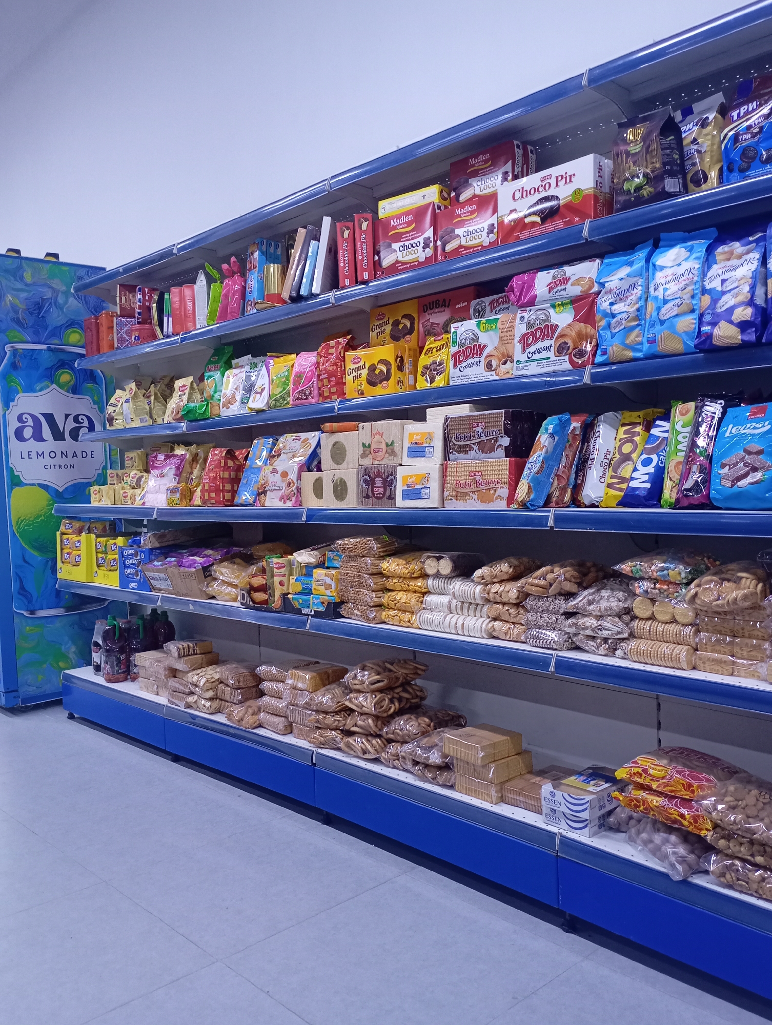 The image shows a well-stocked aisle in a supermarket or convenience store, specifically in the city of Arys, Kazakhstan. The shelves are filled with a variety of packaged food items, primarily sweets and snacks. Dominating the left side of the image is a large blue display panel featuring an advertisement for "Ava Lemonade Citron."
The shelves themselves are light blue and organized with numerous products. The top shelf displays boxes of cookies and chocolates, including brands like "Madlen Fabrica" and "Choco Pir." The second shelf down contains more packaged cookies and snacks, with colorful bags and boxes visible, such as "Dubai" and "Today Croissant" products, as well as several blue bags labeled with Russian text. The third shelf features more chocolate bars and cookies, including "Petit Beurre" and a product labeled "Gorilla." The bottom shelf displays an assortment of bagged snacks, cookies, and some bottled beverages on the far left. The floor is tiled with light gray squares. There are no people visible in the image. The overall impression is one of a typical retail environment.