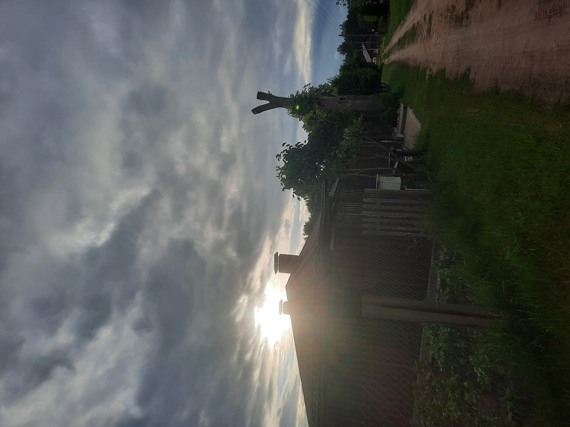 A view from a dirt road in a rural area, likely the village of Porechye, Belarus. The sky is overcast with dramatic grey clouds, but a sliver of bright sunlight breaks through in the upper left, casting a lens flare across the roof of a nearby house. The house is dark-colored with two chimneys. To the right of the house is a tall, bare tree trunk with branches that have been cut, forming a Y-shape. A bright green object, possibly a ball, is attached to the trunk. The road is unpaved, with dry soil and some scattered vegetation. On the left side of the road, there is a chain-link fence enclosing a garden with green plants. Further down the road, several houses with dark roofs are visible in the distance. The scene appears to be captured in the late afternoon or early evening.