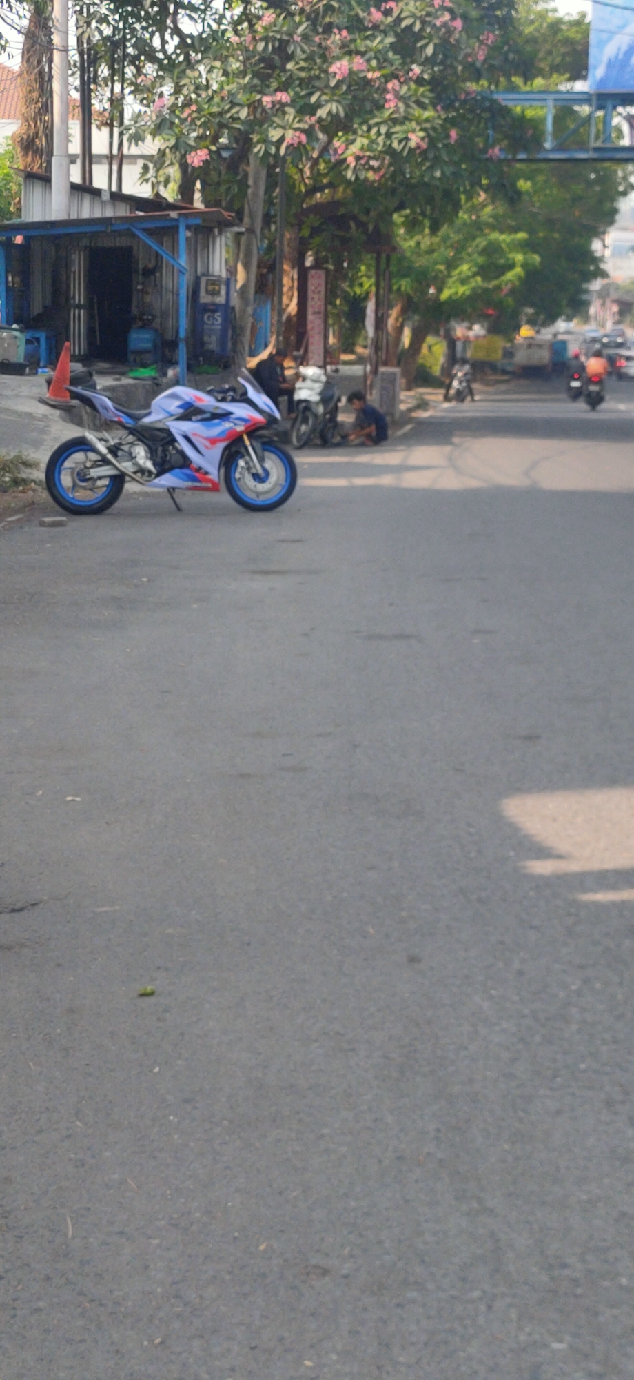 A white, blue, and red sport motorcycle with a flag is parked on the side of a paved road in Semarang, Indonesia. A small open-air shop with a blue roof and shelves is visible behind the motorcycle. Two individuals are seated near another motorcycle in front of the shop. Trees with pink flowers line the area, and traffic can be seen in the background. The road appears to be a common urban street with some debris on the pavement.