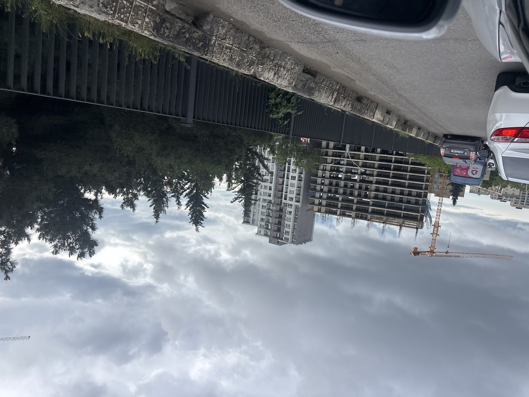 The image depicts a view from a vehicle on a street in Tbilisi, Georgia, facing an urban construction site. A white building is already built, and next to it, an unfinished high-rise under construction with scaffolding and a yellow construction crane extending across the frame. The sky is overcast with heavy clouds. A black metal fence and vegetation are visible on the near side of the road. Several cars are parked in front of the vehicle, including a light-colored sedan and a dark-colored compact car. A portion of the car's side mirror is visible in the upper right corner.