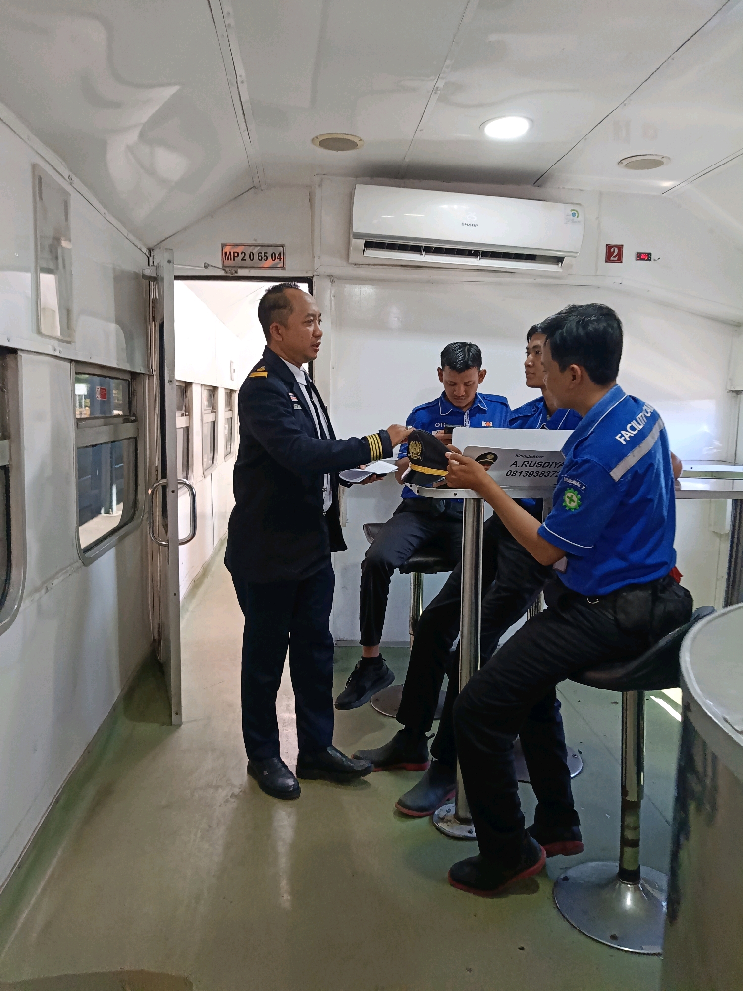 Inside a train car, a man in a dark uniform with gold epaulets stands and gestures while holding papers. He is facing three young men in blue shirts with "FACILITATOR" written on the back. One of the men in blue is holding a cap. The scene appears to be in Surabaya, Indonesia, possibly related to train operations or services. The train car has white walls and ceiling, with windows on the left and an air conditioning unit above. There are tables and chairs present.