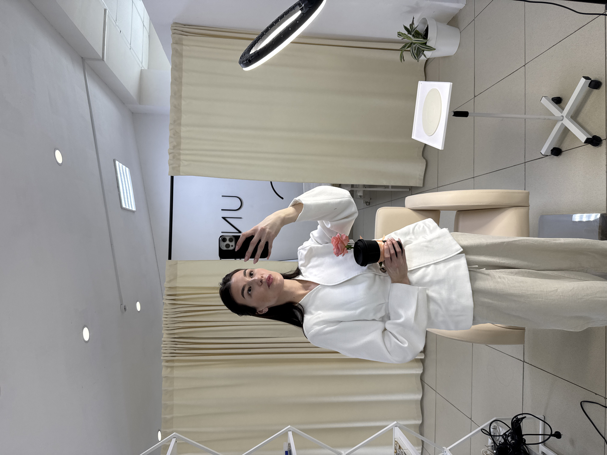 A woman is taking a selfie in a room with beige-toned decor. She is wearing a white jacket and light-colored pants, holding a small black pot with a pink flower. The room includes a ring light, a plant in a white pot, a director's chair, and white shelves. A beige curtain hangs in the background, and "UNI" is visible in reverse on the wall. The location is in the city of Chaikovsky, Russia.