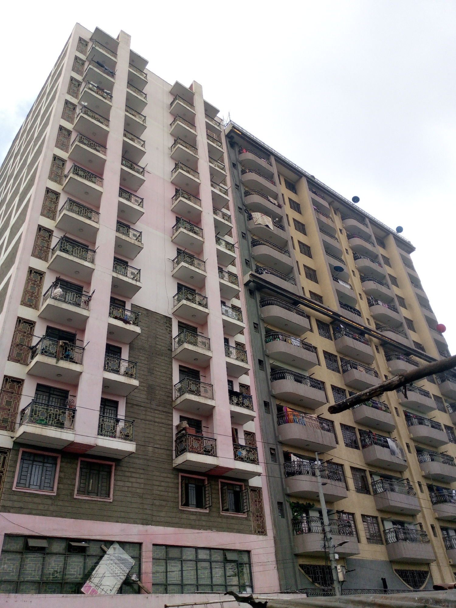The image captures two high-rise apartment buildings in Nairobi, Kenya, viewed from a low angle. The building on the left is primarily pink with a darker, textured lower section, featuring rows of balconies with black railings. The building on the right is beige, also with balconies, some of which have clothing or other items hanging. Several satellite dishes are visible on the roof of the beige building. Overhead utility poles and wires are visible in the foreground. A partially destroyed poster obscures the ground floor of the left building. The sky is overcast.