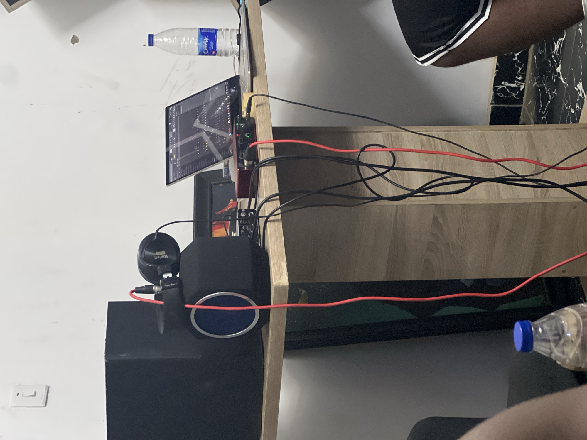 The image shows a section of a desk or shelving unit against a white wall. Objects on the desk include a computer monitor, audio interface, microphone with pop filter and headphones, and multiple power or audio cables. There are two water bottles visible. The environment includes a light switch on the white wall. Part of a person's leg is visible. I am unable to confirm the location in the city of Abuja, Nigeria.