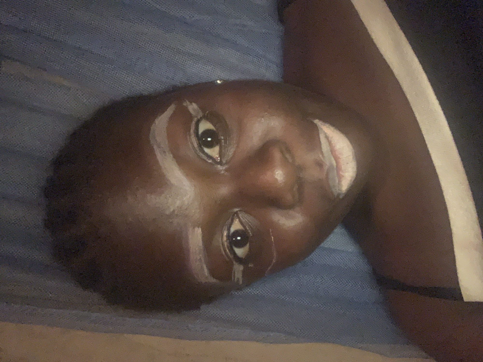 The image presents a close-up, rotated view of a dark-skinned individual, likely a child or young person, lying down and looking directly at the camera. The subject's face is adorned with prominent white markings: lines of a white substance are drawn above and below both eyes, extending onto the forehead in an arc, and also covering the area around the mouth. The eyes are wide open, engaging the viewer.
The person appears to be resting on a blue, textured fabric, which resembles a mosquito net or a patterned blanket, indicating an indoor setting. On the right side of the frame, a dark garment with a distinct white stripe is visible. The overall lighting is dim, suggesting that the photograph was taken in low light conditions, possibly during the evening or night. There is no visible text in the image.
Given the context of Kukum, Nigeria, the white facial markings are highly indicative of a traditional or cultural practice. Such body art is common in many Nigerian cultures for ceremonies, rituals, festivals, or as a form of traditional make-up, often using substances like chalk or clay. The image captures a moment of this cultural expression, with the individual presenting themselves directly to the camera.
