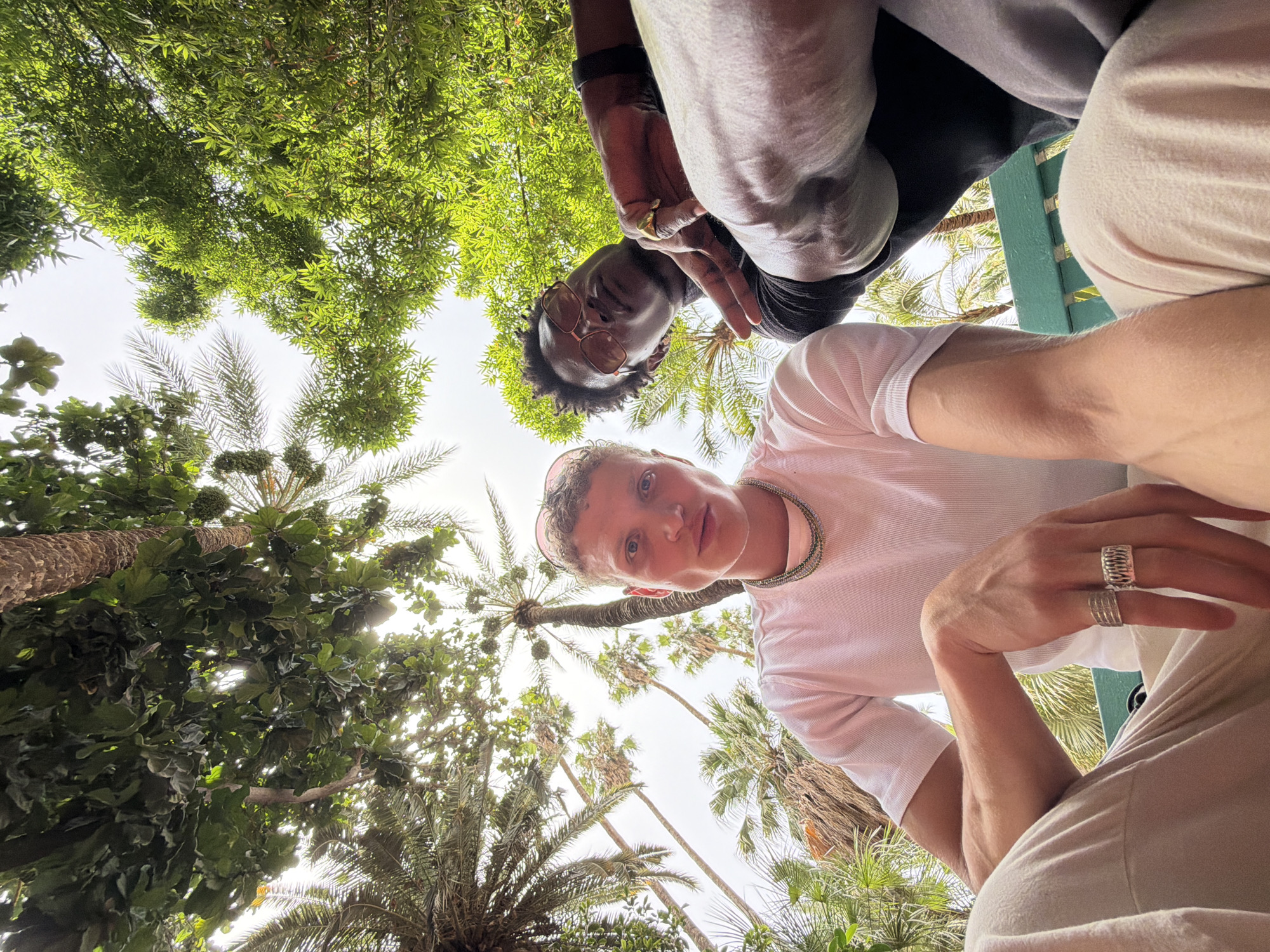 In Marrakesh, Morocco, two individuals are captured from a low-angle perspective beneath the canopy of palm trees and lush green foliage. The man on the left, with fair skin and short, light-colored hair, wears a white t-shirt and a multi-layered necklace. He is adorned with several silver rings on his left hand. The man on the right has darker skin and is wearing sunglasses and a black t-shirt. He has a gold ring on his right hand and is making a peace sign with his left hand. Both are seated on a green bench. The scene is characterized by the interplay of sunlight filtering through the dense greenery, creating a natural, outdoor setting.