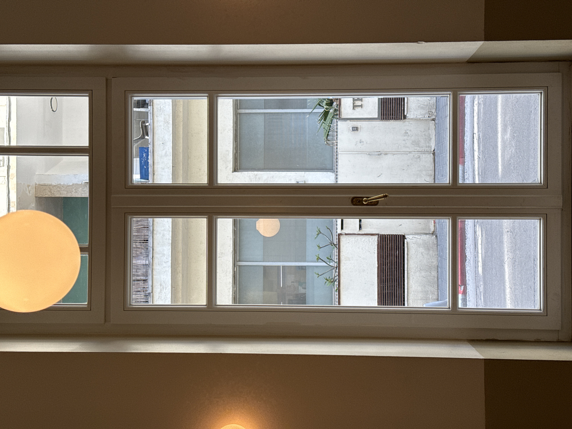 The image shows a window looking out onto a street in Tel-Aviv, Israel. Through the window, buildings with varied architectural styles are visible, including concrete structures and facades with vertical elements. A small tree or bush grows outside one of the windows. A street with a red curb is visible. The window itself is white with a horizontal divider. Two light fixtures hang on the inside, one a round yellow glow on the left and the other a small glow below the windowsill.