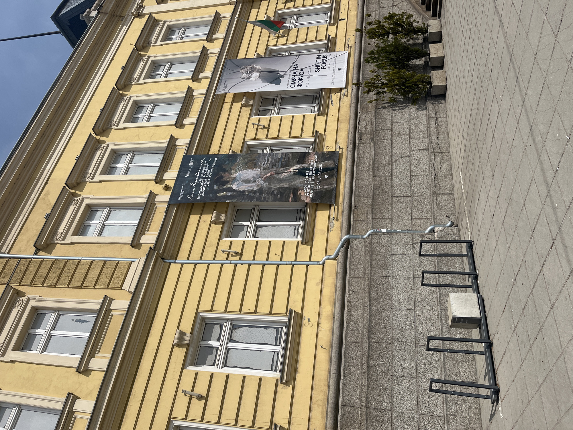 The photograph captures a building facade in Sofia, Bulgaria, adorned with multiple exhibition banners. The building has a yellow facade with decorative window frames. Three prominent banners are hung on the building. The top two banners advertise an exhibition titled "Shift in Focus" by Mirena Zlateva, running from 21.01-12.06.2026. The lower banner commemorates the 150th anniversary of Elena Karamikhailova's birth. A Bulgarian flag is visible near the top. A gutter runs along the side of the building. A gray sidewalk with tiled surfaces is to the side of the building, complete with a black metal bicycle rack.