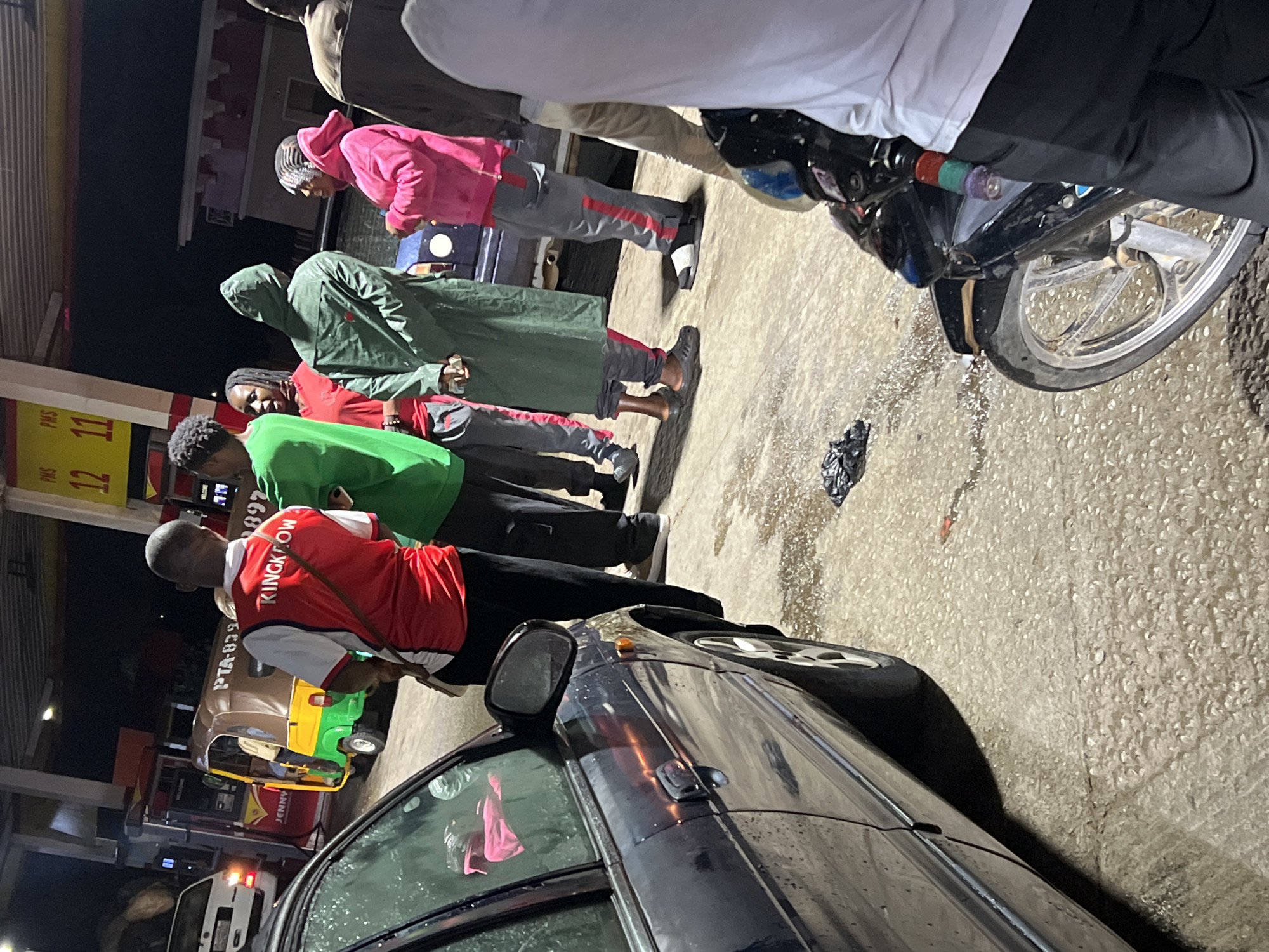 The image depicts a nighttime scene at a gas station in Gura Top, Nigeria, where several individuals are gathered, potentially waiting for fuel or service. The ground is wet, suggesting either recent rain or liquid spillage.
In the foreground, a dark sedan is partially visible on the left. To its right, a person wearing a red vest with "RINGKOW" written on the back, over a white t-shirt, stands facing away from the camera. This individual is positioned near a line of people. Behind them are several other individuals: one in a green t-shirt and dark pants, another in a red top and dark pants, a person in a green hooded jacket and grey sweatpants with red stripes, and a person in a pink hooded top and similar grey sweatpants. A partially visible person in a white shirt stands next to a black motorcycle on the far right, which has colorful handle grips.
A gas pump with yellow and red signage is visible in the background, displaying "PMS" along with the numbers "12" and "11". A brown, yellow, and green tricycle, commonly known as a keke napep, is parked behind the line of people, with partial text "PTA-..." visible on its side. Another red structure in the background shows a partially visible word, "JENNY". The scene is illuminated by artificial lights against the dark night sky.