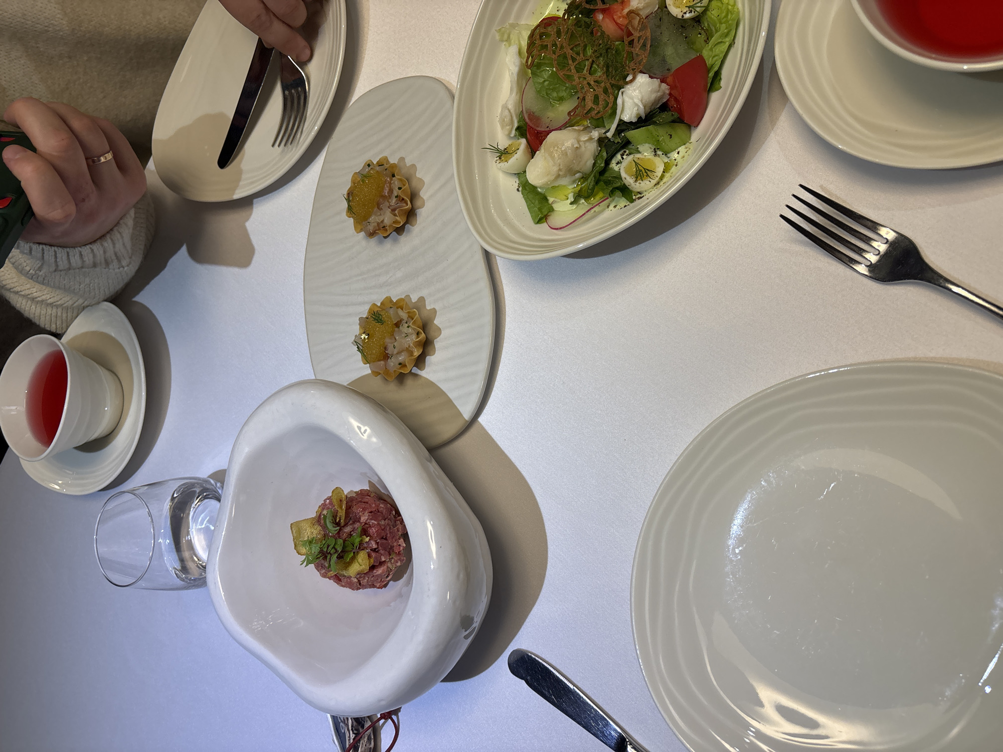The image depicts an indoor dining setting at a restaurant. A white tablecloth covers the table. In the upper right quadrant, a person's hand, adorned with a gold-colored ring on the ring finger, holds a dark green smartphone. Adjacent to this, a white teacup containing a red-pink liquid rests on a saucer. Directly below, another hand holds a silver fork and knife over an empty oval-shaped white plate.
Prominently displayed on the table are several dishes. In the center-left, an irregular, organic-shaped white bowl contains a mound of finely minced raw meat, likely beef tartare, garnished with small green herbs and yellow crisps. To the right of this, on a rectangular white plate with subtle ridged patterns, are two small, fluted pastry cups (tartlets). Each tartlet contains a light-colored, diced filling topped with orange roe and a sprig of green dill.
In the lower right quadrant, a large, oval-shaped white bowl holds a colorful salad. Visible components include green leafy lettuce, red tomato wedges, thin slices of radish, white pieces of what appears to be cheese (e.g., mozzarella), and halved quail eggs. A delicate, intricate, golden-brown crisp network garnishes the salad, which also features a light dressing and small green herbs, possibly dill.
Other objects on the table include a clear glass containing water, an empty round white plate, and a silver fork positioned near the bottom center. The specific location within Krasnoyarsk, Russia, cannot be determined from the visual evidence presented in the image. The environment is consistent with a modern, upscale dining establishment.