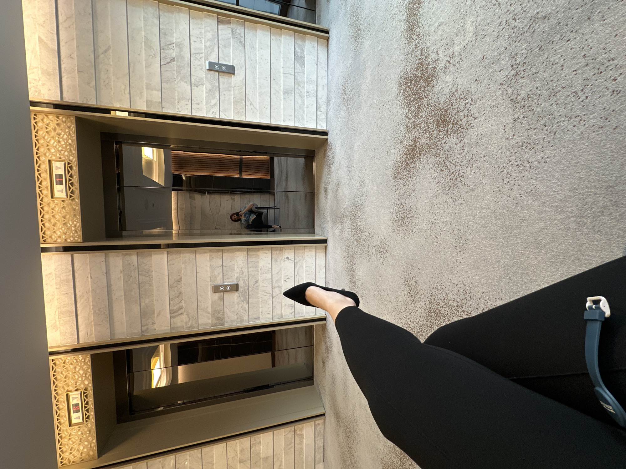 The image depicts an interior space, possibly a high-end lobby or residential building in Kazanlak, Bulgaria. Two elevators are visible. One contains a person sitting on the floor. The foreground includes a person's legs in black pants and a black pointed shoe. A watch with a blue band is visible on the right leg. The floor is carpeted with a speckled, light brown pattern. The elevator exteriors have marble-like panels and decorative metallic elements around the doors. Elevator buttons are present.