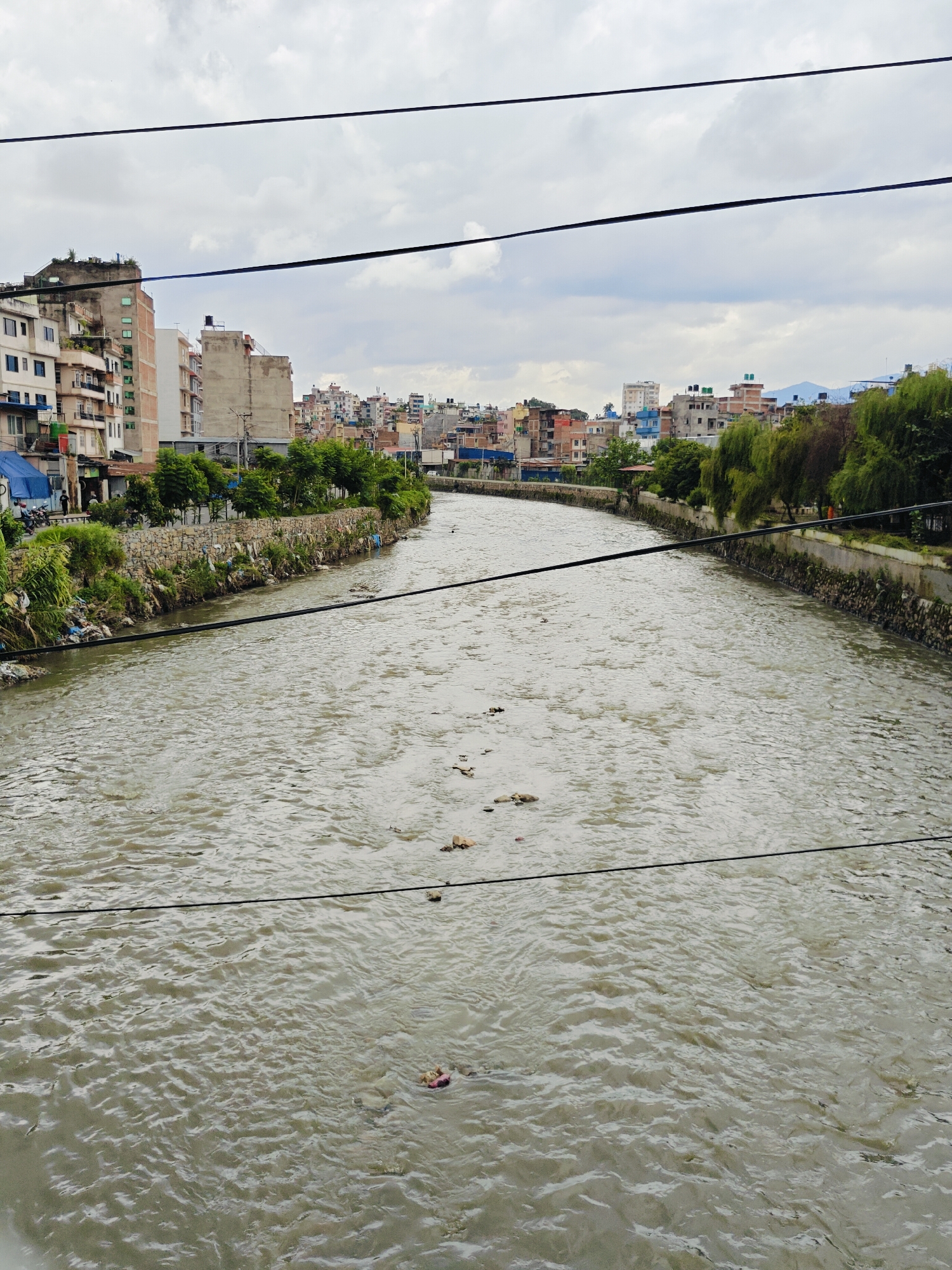 The image shows the Bagmati River in Kathmandu Metropolitan City, Nepal. The river is wide and murky, with visible debris floating on the surface. The banks of the river are lined with stone walls, and vegetation grows along the edges. In the background, a dense urban landscape with multi-story buildings is visible. The sky is overcast with clouds. Overhead, power lines stretch across the frame. The scene depicts a polluted urban river within a bustling city.