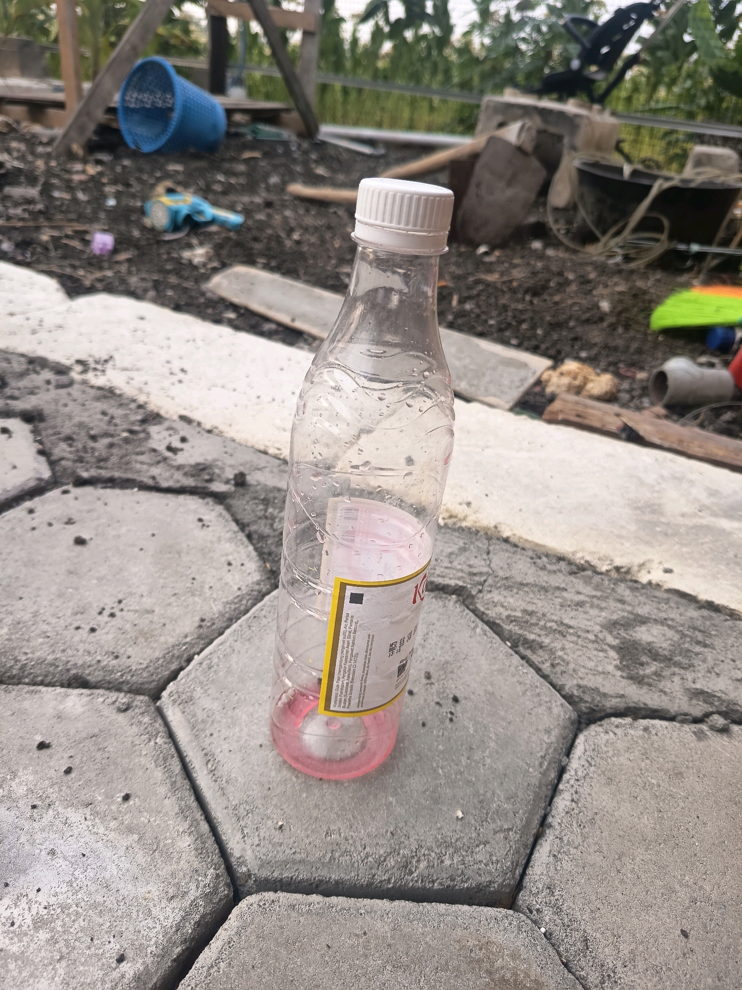 In Sidorejo, Indonesia, a clear plastic bottle with a white cap sits on a hexagonal paved surface. The bottle is half-full of a pink liquid and has a yellow label. The background shows an outdoor area with a blue basket, some debris, and a wooden structure. The ground is a mix of dirt and light-colored paving stones, with some green vegetation visible in the distance.