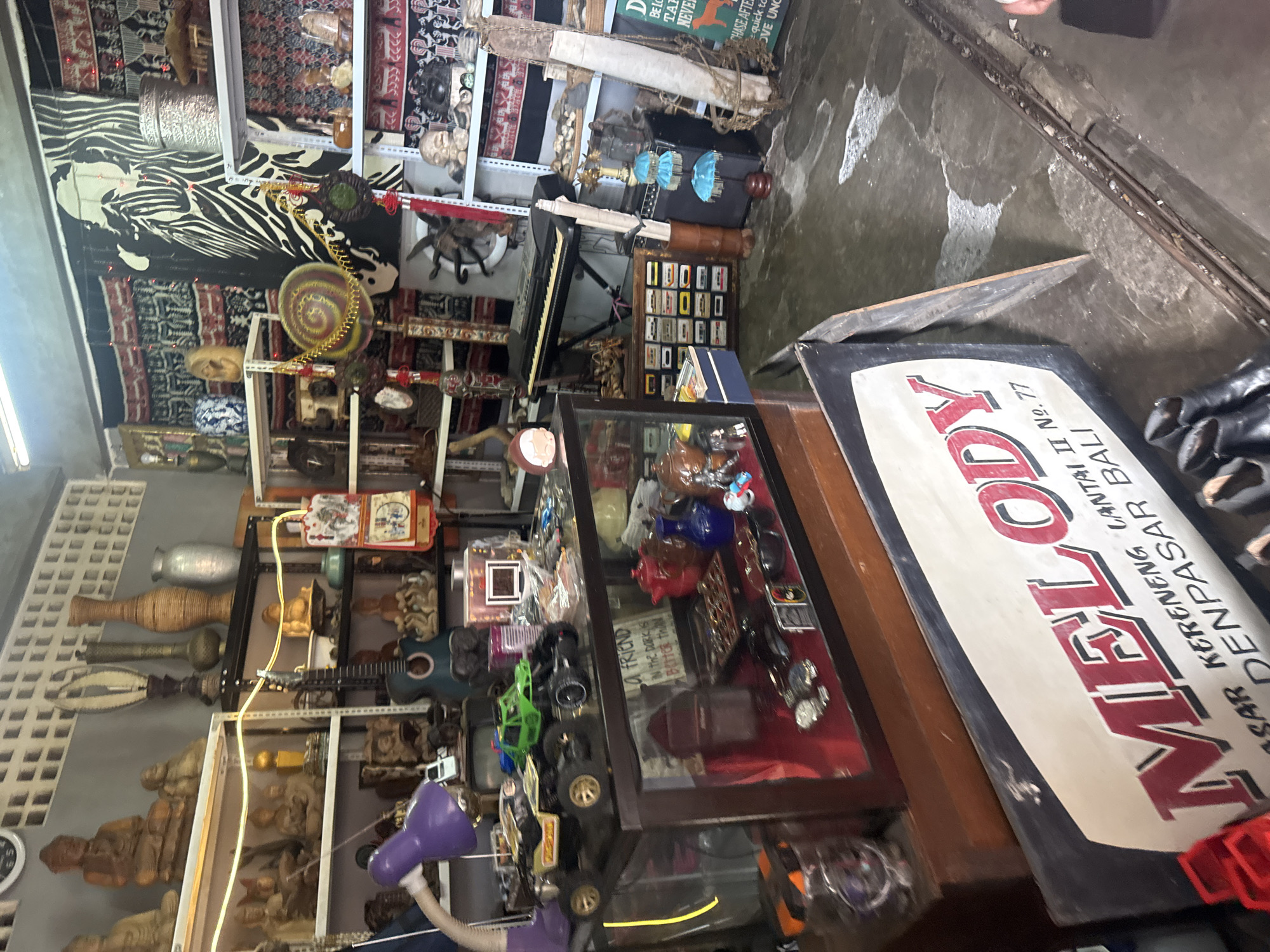 The image depicts the interior of a shop in Denpasar, Indonesia, likely a souvenir or antique store given the eclectic mix of items. The shop is densely packed with various objects arranged on shelves, displayed in glass cases, and placed on the floor.
In the foreground, a large, weathered sign with red lettering reads "MELODY" with smaller text underneath in Indonesian, indicating the shop's name and location. To the left of the sign are some red plastic items.
The shop's interior features numerous shelves stocked with a variety of items. On the top shelves, various statues, vases, and decorative objects are visible, including what appear to be religious figurines and traditional Indonesian crafts. There is also a keyboard on a stand in the middle of the shop, suggesting a possible musical element to the store.
A prominent glass display case in the center of the image showcases smaller items such as toy cars, sunglasses, watches, and other trinkets. On the shelves above and behind the display case, more items are visible, including a guitar, various decorative pieces, and what looks like a calendar or poster.
The walls of the shop are adorned with textiles, including patterned fabrics and a large black and white piece with a striking zebra-like design. The lighting in the shop appears to be a mix of overhead fixtures and string lights, creating a somewhat dim and cluttered atmosphere. The floor is a rough concrete surface, showing signs of wear.
While no individuals are clearly visible and actively engaged in an interaction, the presence of the store and its contents implies that it is a commercial establishment open for business. The overall scene conveys a sense of a well-trafficked, eclectic marketplace filled with a diverse array of goods.