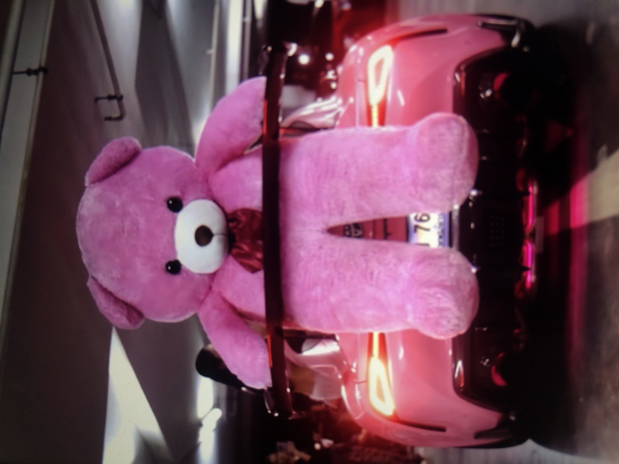 The image shows a large pink teddy bear strapped into the driver's seat of a pink car. The car's headlights are on. The location is unknown, and there is no indication in the image that it is in Lhokseumawe, Indonesia.