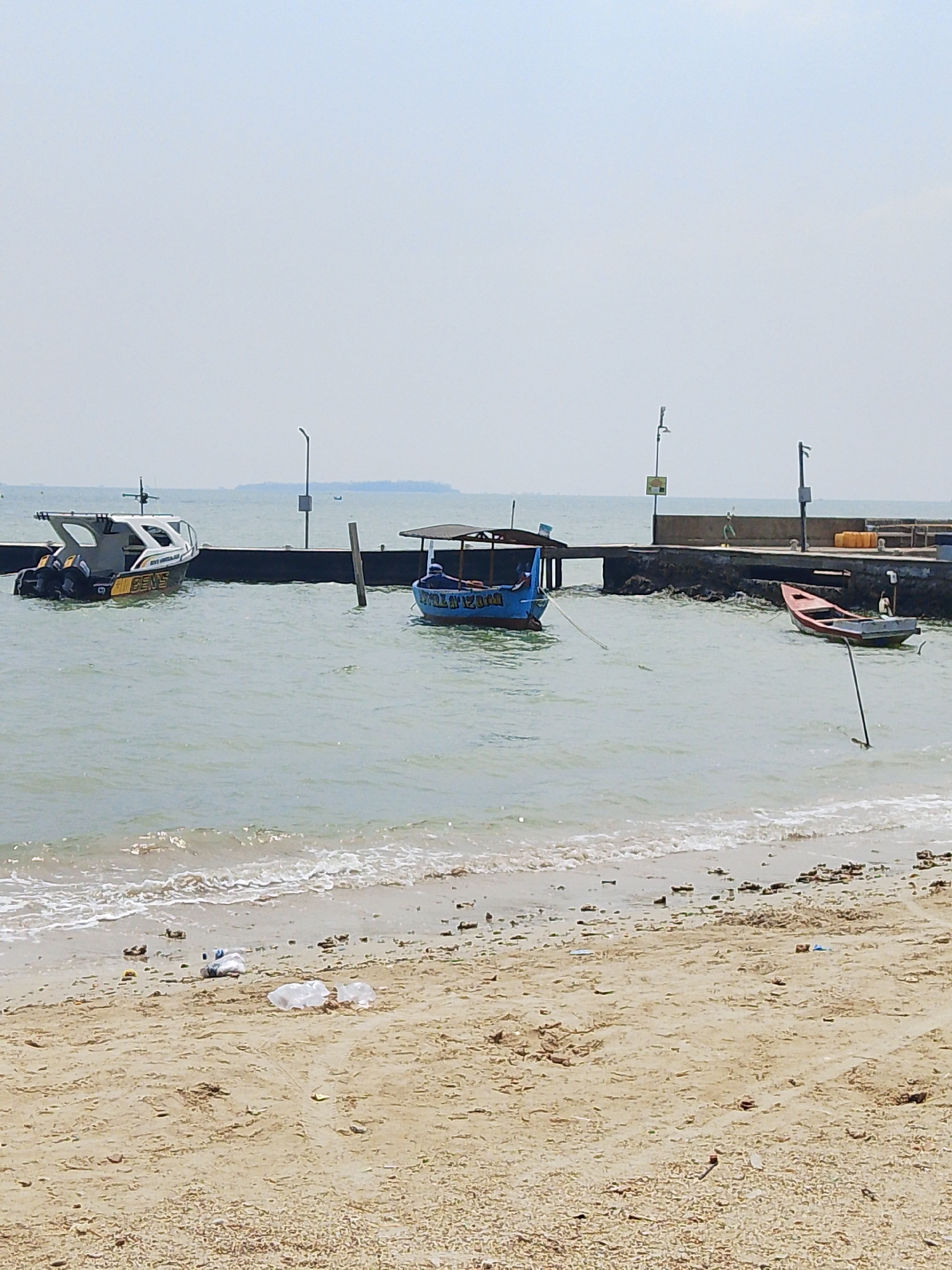 The image captures a section of the coastline in Pejagan, Indonesia, with a sandy beach in the foreground and the sea beyond. A concrete pier extends into the water, with several boats moored alongside. Two of the boats are small fishing vessels, one blue and the other red, while a larger, white and grey boat with outboard motors appears to be a patrol or utility craft. The water is calm with small waves lapping at the shore. Debris, including plastic bottles and other refuse, is scattered on the beach. The sky is overcast, suggesting a hazy or cloudy day.