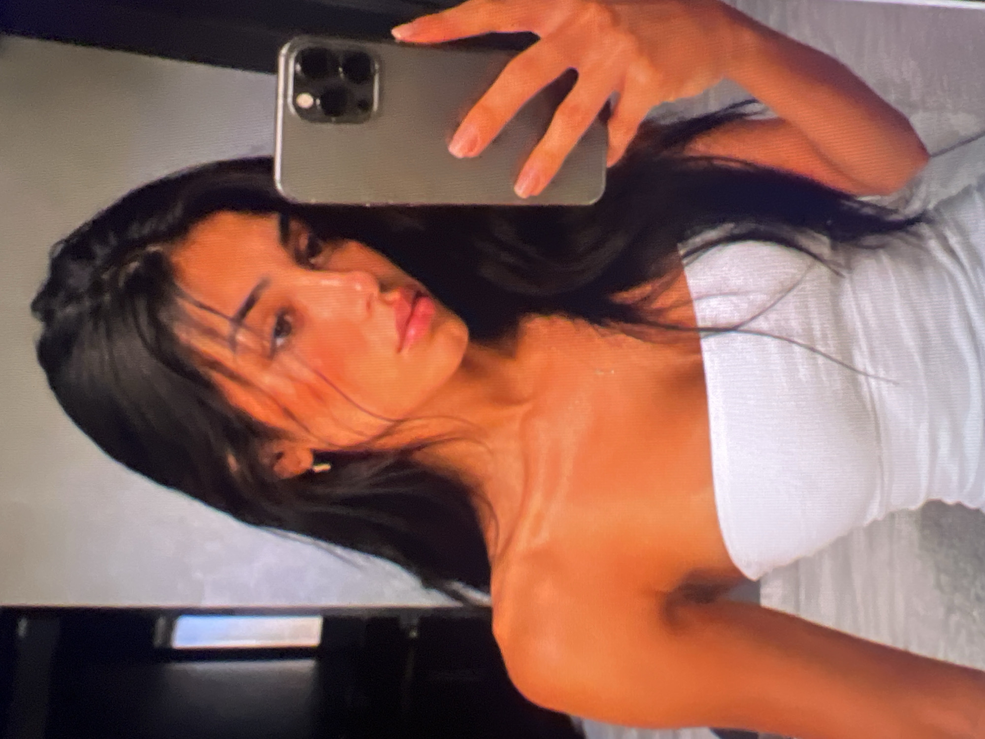 A young woman with long, dark hair is taking a mirror selfie. She is wearing a white strapless top. She is holding a dark-colored smartphone in her right hand, which is positioned to capture the reflection. Her left hand is reaching up to touch her hair. The background appears to be an indoor setting, possibly a bedroom or bathroom, with a neutral-colored wall and a dark vertical element on the left side of the frame. The lighting is warm, casting an orange hue across the scene.