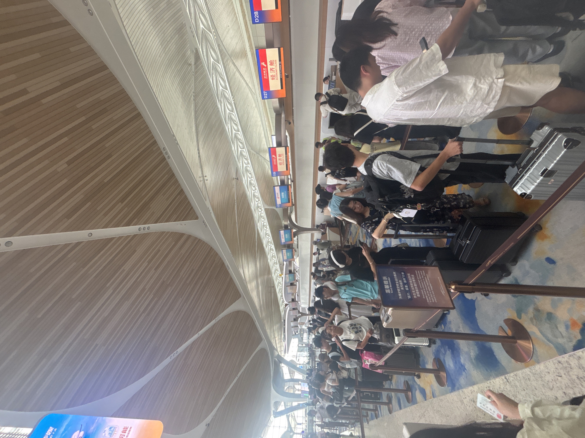 The image shows a busy airport check-in area, likely in Baiyun District, China, indicated by the presence of Chinese text on signs and digital displays. A long queue of people, mostly appearing to be tourists, are lined up at several check-in counters. They are carrying luggage, including suitcases and backpacks, and some are using their mobile phones. The check-in counters have digital displays showing flight information and gate numbers, with one display clearly indicating "ECONOMY CLASS" in both English and Chinese. The environment features a modern architectural design with a wooden slatted ceiling and white structural elements. The floor is covered with a colorful carpet. Barriers and stanchions guide the queue. The lighting is bright, suggesting it is daytime.