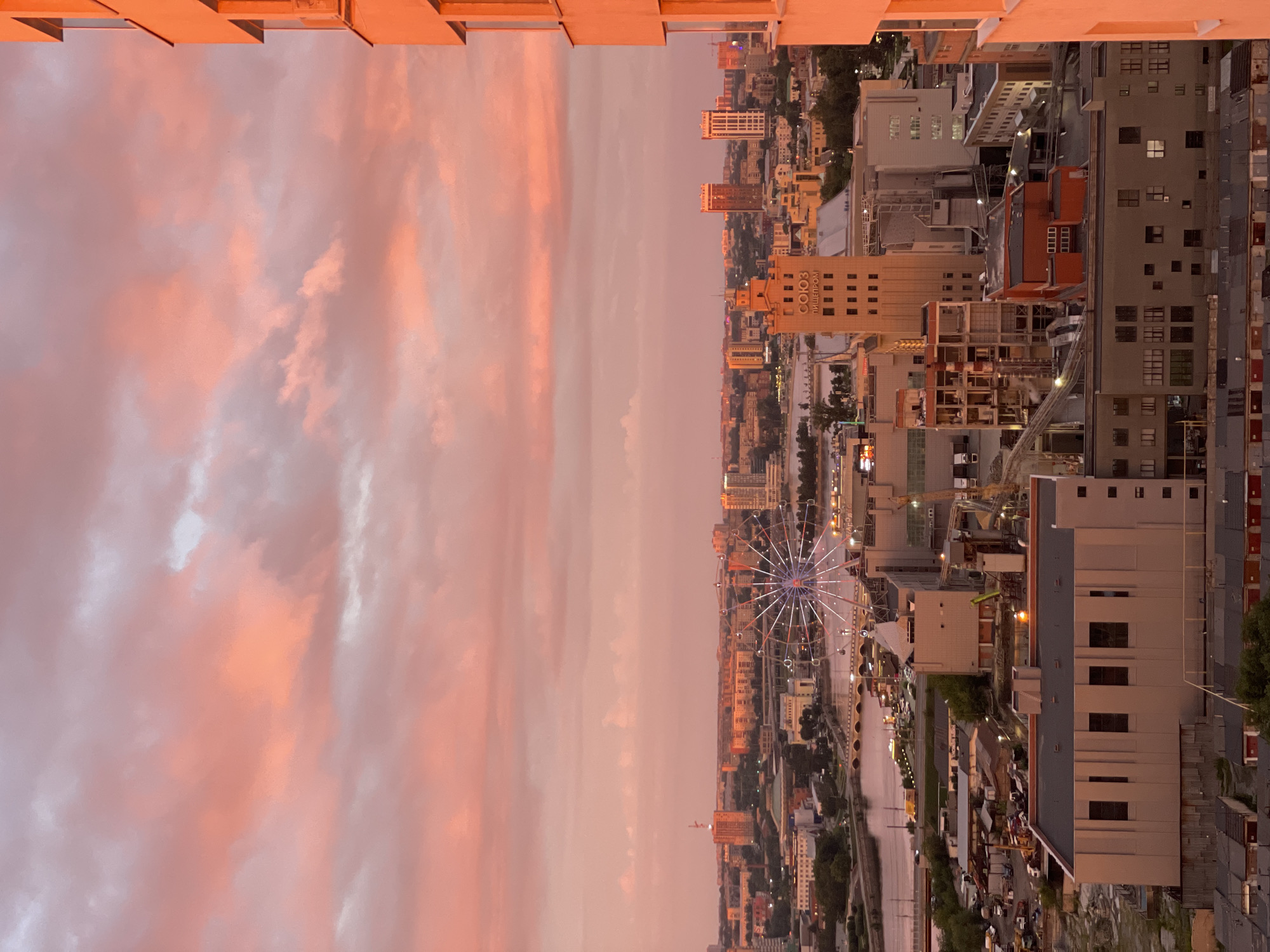 This is a vertical, high-angle shot of a city at dusk. The sky is filled with dramatic, sunset-colored clouds, ranging from pale pink to deeper orange. The cityscape to the right is illuminated by the fading light, with buildings of various heights and architectural styles. A prominent Ferris wheel, lit up with colorful lights, stands out in the middle distance. A river or canal runs through the city, reflecting the sky. The scene conveys a sense of urban beauty at the end of the day. Some visible text on a building reads "СОЮЗ ПИЩПРОМ".