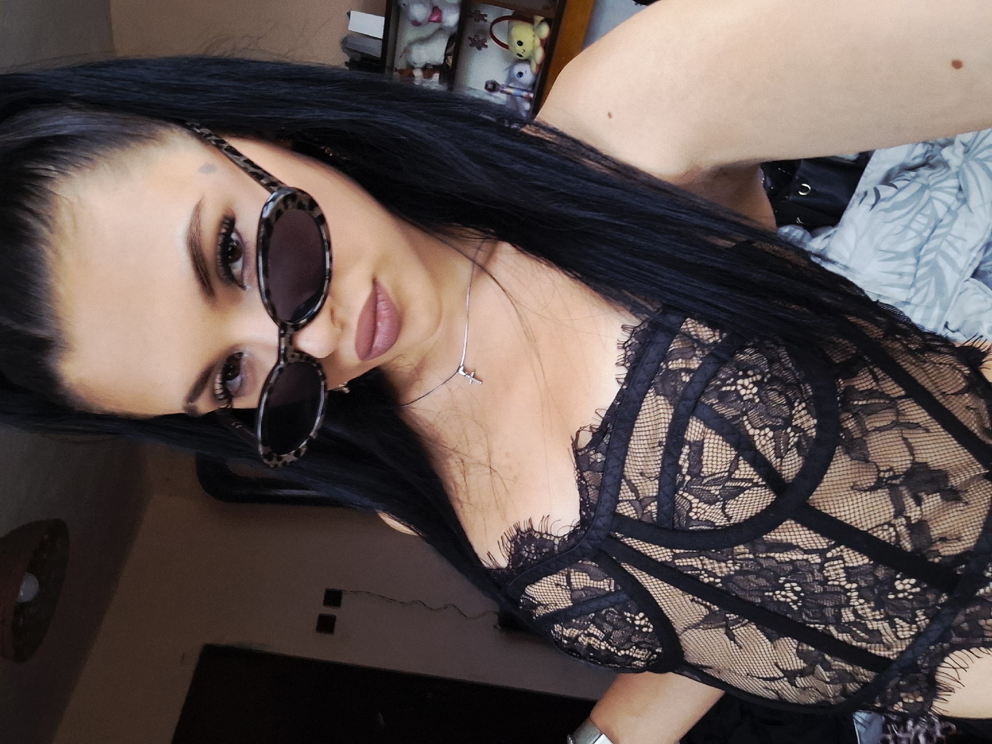 A young woman with long, straight, dark hair takes a close-up selfie in an indoor setting. She is positioned diagonally in the frame, looking towards the camera with a subtle expression. Her eyes are partially obscured by stylish leopard-print framed sunglasses with dark lenses. She wears matte nude-pink lipstick. Around her neck is a silver chain necklace featuring a small, airplane-shaped pendant. She is dressed in a black lace and mesh top or bodysuit, which features intricate floral patterns and opaque strap details over a sheer base. A silver-colored watch or bracelet is visible on her right wrist at the bottom right of the image.
The background suggests a casual, domestic environment, possibly a bedroom or living room. In the upper left, blurred shelves display small decorative items, including what appear to be stuffed animals (a yellow bear and another light-colored one). To the upper right, a light-colored fabric with a subtle leaf pattern is visible, possibly a blanket or bedding. The lower left shows a darker wall with a circular light fixture. The overall lighting is soft, suggesting daytime indoors. No visible text is present in the image. The scene takes place in Krapets, Bulgaria.