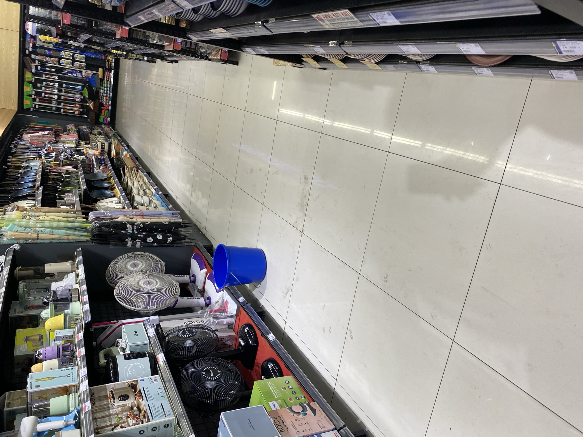 The image shows a bright, well-lit aisle in a retail store, likely a supermarket or a department store, located in Banda Aceh, Indonesia. The floor is made of large, pale tiles, with faint smudges and scuff marks visible, indicating it is a high-traffic area. On the left side of the aisle, shelves are stocked with various household appliances. This includes blenders, hand mixers, and electric fans. A prominent blue plastic bucket sits on the floor near the shelves. To the right, more shelves display kitchenware, including plates and bowls. The aisle extends into the distance, with reflections from overhead lighting visible on the floor. The overall impression is of a clean, organized retail environment.