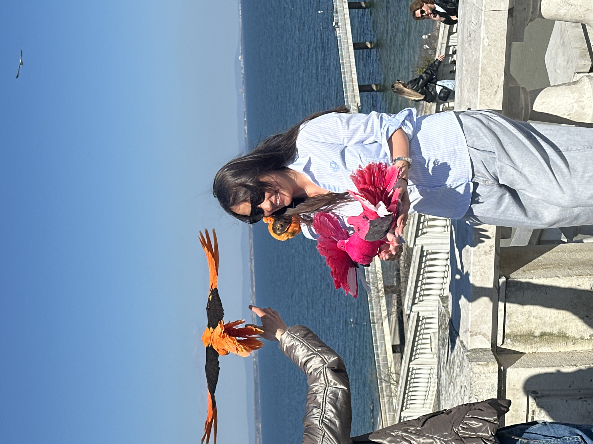 A woman with long dark hair wearing sunglasses and a light blue striped shirt is holding a bouquet of colorful artificial flowers. Another person's arm is extended towards a large artificial bird with black body and orange wings, seemingly releasing it into the air. The background shows a body of water, a pier, and people sitting on a ledge. The photo appears to be taken outdoors on a sunny day. The specific location of Burgas, Bulgaria cannot be determined with certainty based on the image alone.