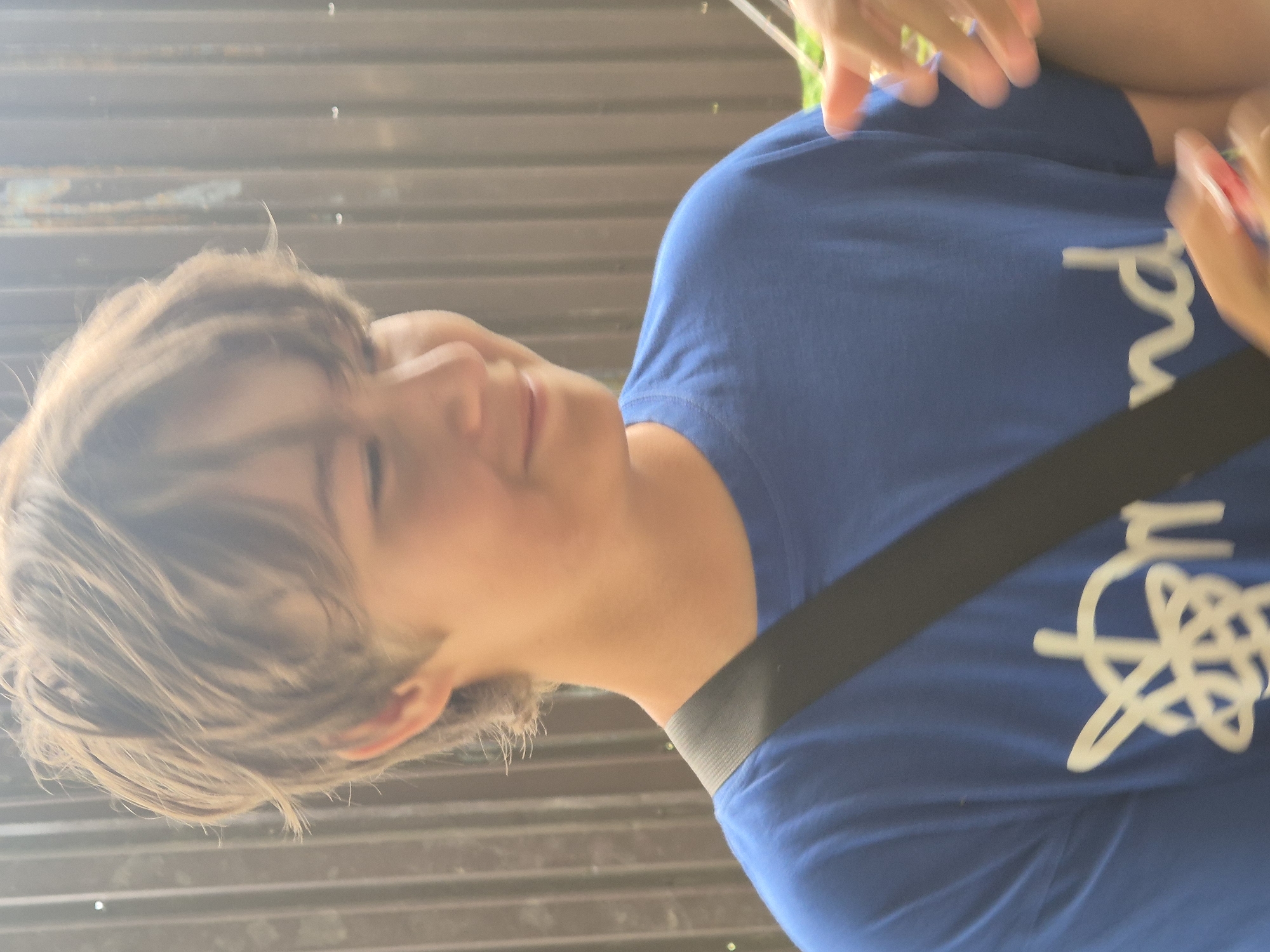 A young boy with light brown hair and a blue t-shirt is pictured outdoors in front of a brown corrugated metal fence. The boy is looking down and to his right, with a slight smile. He has a black strap going across his chest. The t-shirt has a white graphic design on the front, with the letters "nd" visible. The overall lighting suggests it is daytime. The location is Sozonivka, Ukraine.