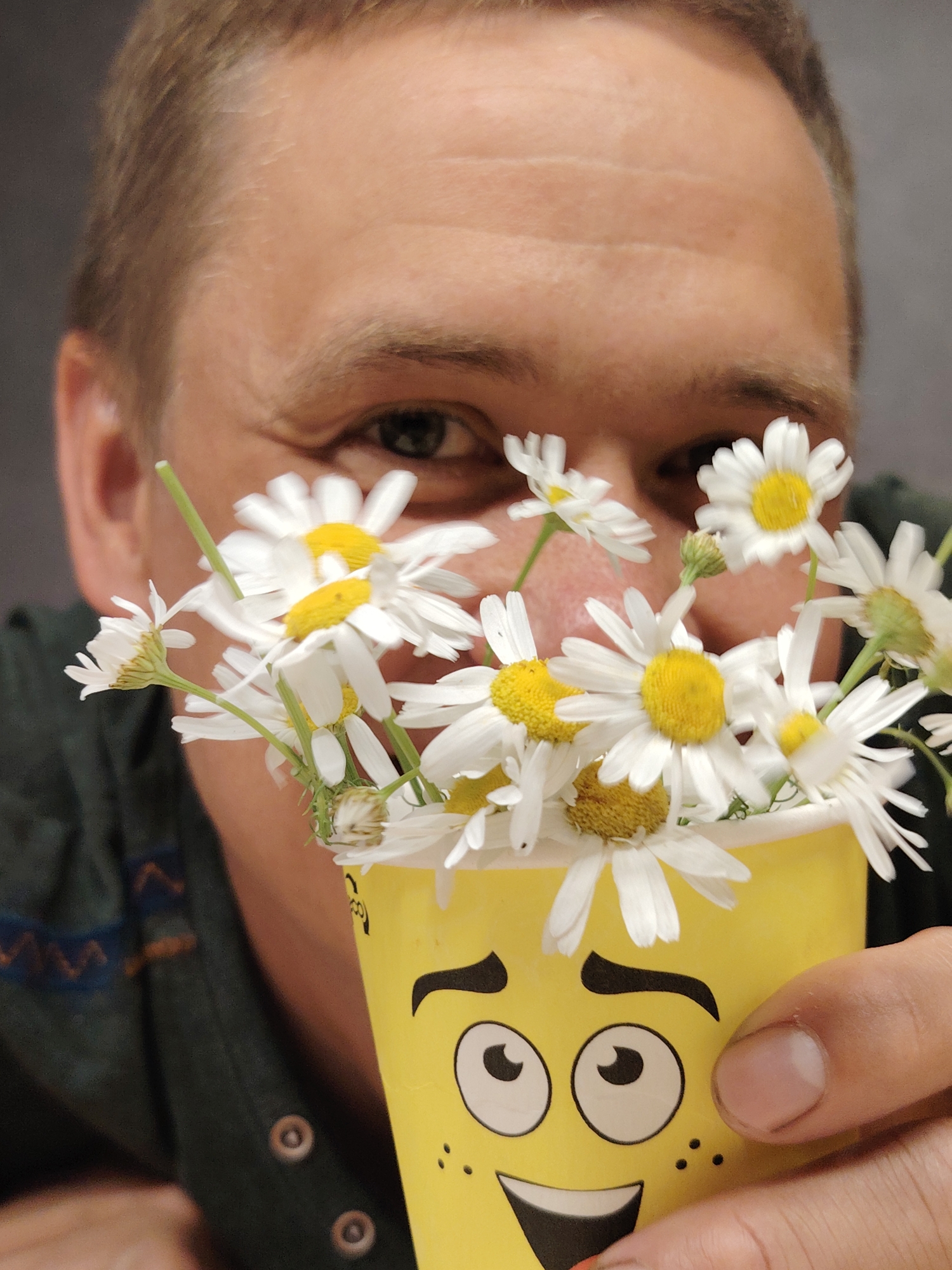 The image shows a close-up of a person holding a yellow cup with a smiley face design. The cup is filled with daisies. The person's face is partially visible behind the flowers, and they appear to be looking at the camera. The background is a plain gray wall. The image does not contain any identifiable landmarks or clues that would suggest a specific location within the city of Chaikovsky, Russia.