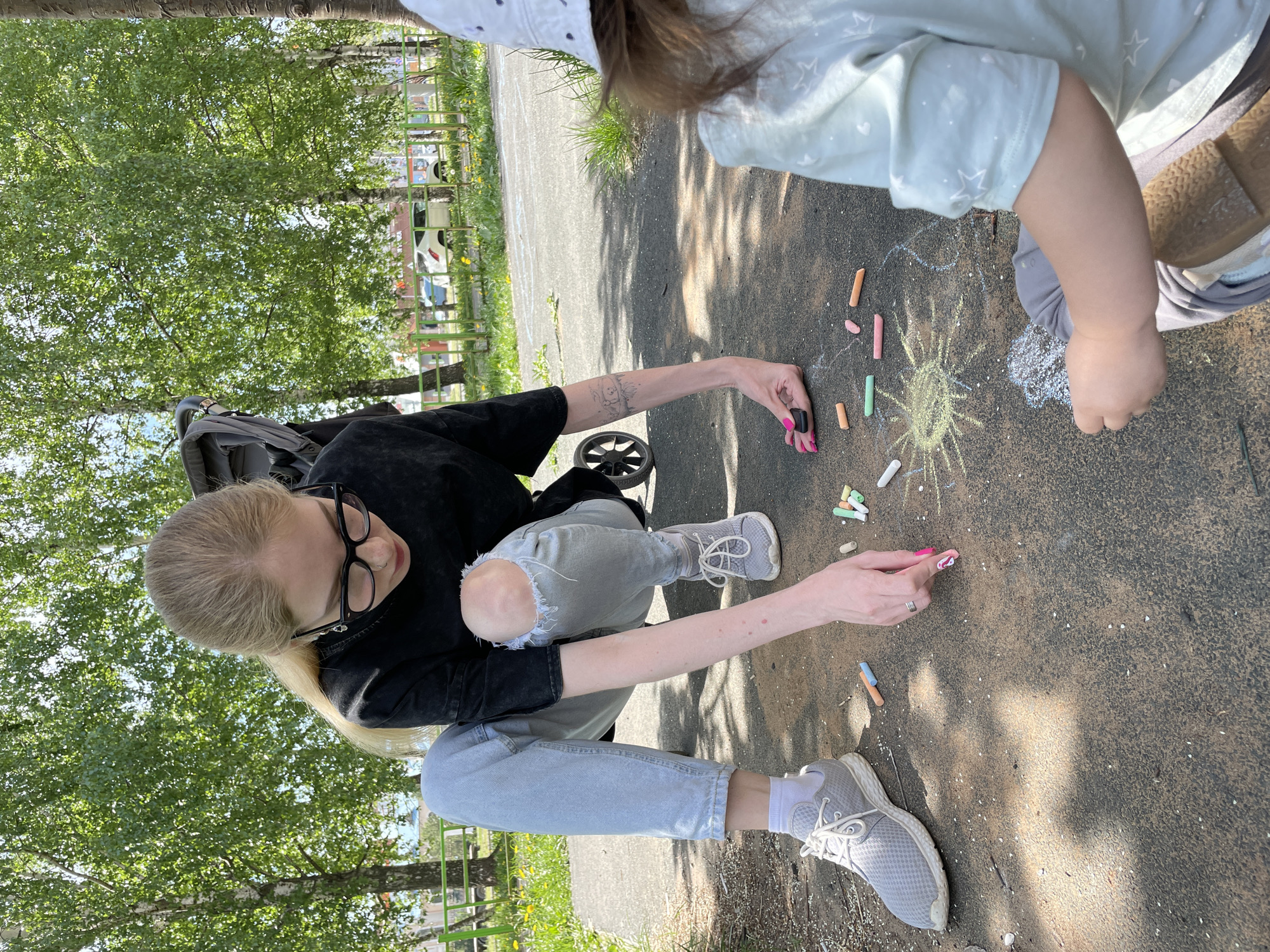 A woman in a black shirt, ripped light-wash jeans, and grey sneakers crouches on the ground, drawing with chalk. Next to her, a small child wearing a light-colored shirt and hat is also drawing. Chalk pieces of various colors are scattered around their drawings. A stroller is behind the woman. The scene is outdoors on a paved surface, possibly a sidewalk, surrounded by trees and grass. A green fence is visible in the background. The photo was taken in Izhevsk, Russia.