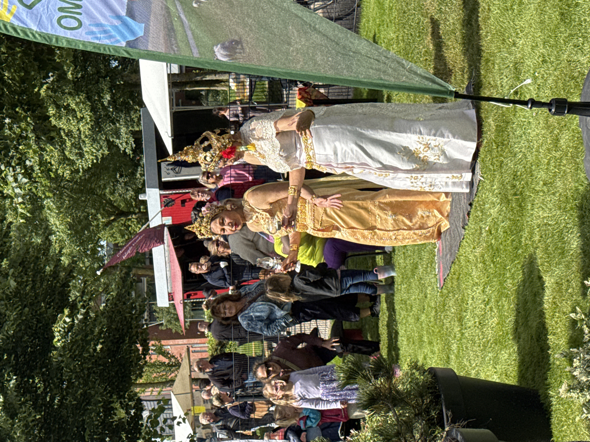 In an outdoor setting with a grassy lawn and trees, two women in traditional Southeast Asian attire, featuring elaborate headdresses and golden ornamentation, are posing. The woman on the left wears a shimmering golden garment, while the woman on the right is dressed in a white gown with gold embroidery. Both are holding what appear to be glasses, possibly containing beverages. Several other people, including adults and children, are visible in the background, suggesting an event or gathering. Behind them, there are stalls or stands, indicating a public space such as a market or festival. A green mesh flag with text that is partially obscured is positioned in the foreground on the right. The lighting suggests it is daytime.