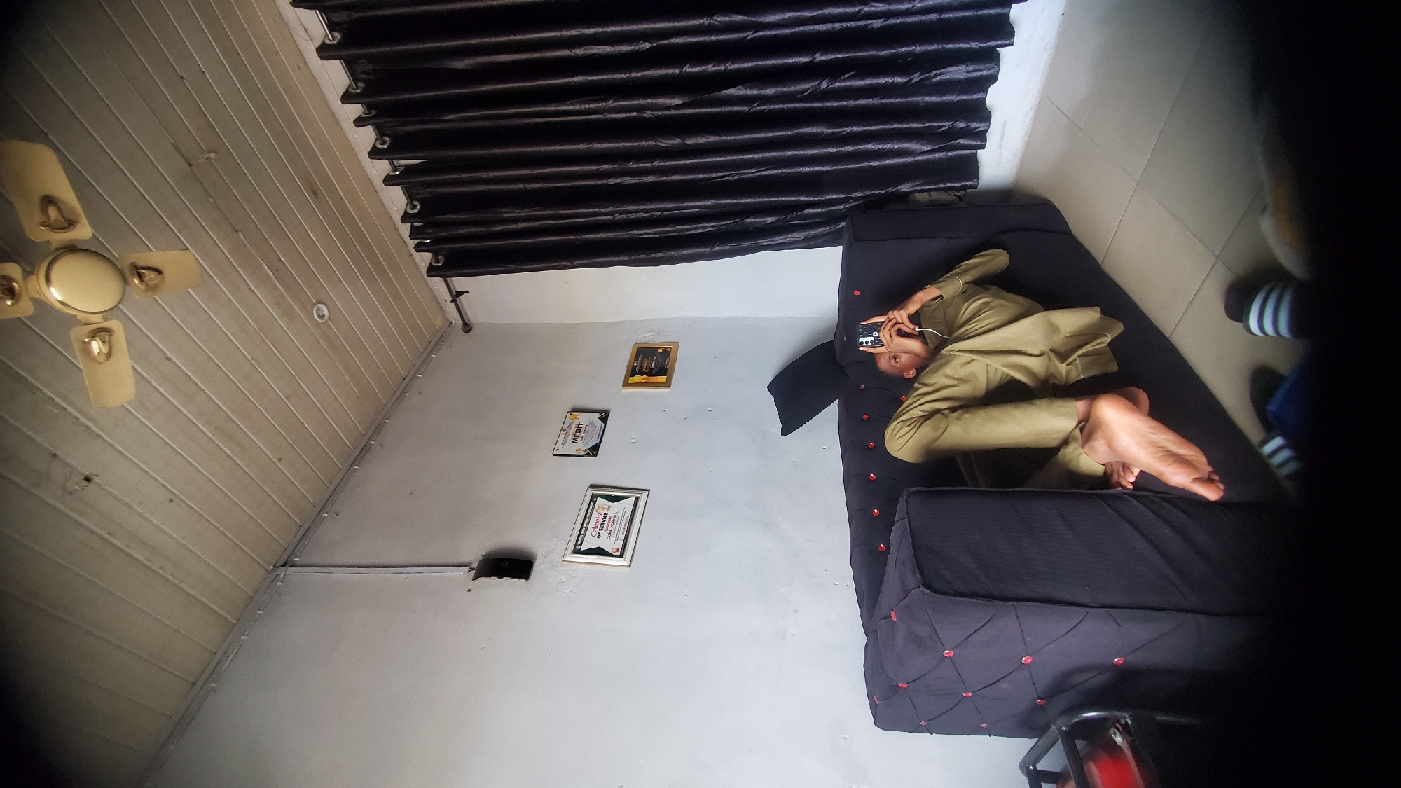 This image depicts an indoor scene in Jalingo, Nigeria, featuring an individual on a sofa within a moderately furnished room.
**People:** One individual, appearing male and young adult, is centrally positioned, lying reclined on their back on a dark upholstered sofa. The individual wears a light brown/khaki-colored long-sleeved tunic and matching trousers. Their bare right foot is prominently visible, angled towards the foreground. They are holding a black smartphone vertically with both hands, looking down at the screen, with a white earbud wire extending towards their right ear. At the very bottom of the frame, two feet, partially obscured, are visible; one wears a striped sock, and the other a dark sock with part of a dark sandal.
**Objects & Furniture:** The primary furniture is a dark grey/black L-shaped or modular sofa, featuring a quilted design with red buttons on the visible armrest/backrest section. A black pillow supports the individual's head. Three framed items are mounted on the light-colored left wall: the leftmost frame contains an "Award of Service" certificate, the middle frame displays a "MERIT" certificate, and the rightmost is a smaller, gold-framed item with illegible text. A dark, rectangular object, possibly a speaker or an electrical fixture, is mounted high on the left wall above the awards. A four-bladed, cream-colored ceiling fan with gold accents is suspended from the ceiling. Dark brown/black pleated curtains cover a window or doorway on the right side of the room, hung on a thin metal rod. A partial view of a dark, metal-framed side table with a red object on top is visible in the lower left corner.
**Environment:** The room features light-colored, possibly off-white or light grey, textured walls showing some wear. The ceiling is constructed from light-colored PVC paneling, laid horizontally, with visible seams. Grey electrical conduits run along the wall-ceiling junctions and vertically down the left wall. The floor is covered with light-toned square ceramic tiles with dark grout lines. The overall lighting appears to be a mix of natural light from behind the curtains and interior ambient light, possibly from a small recessed light fixture visible in the ceiling.
