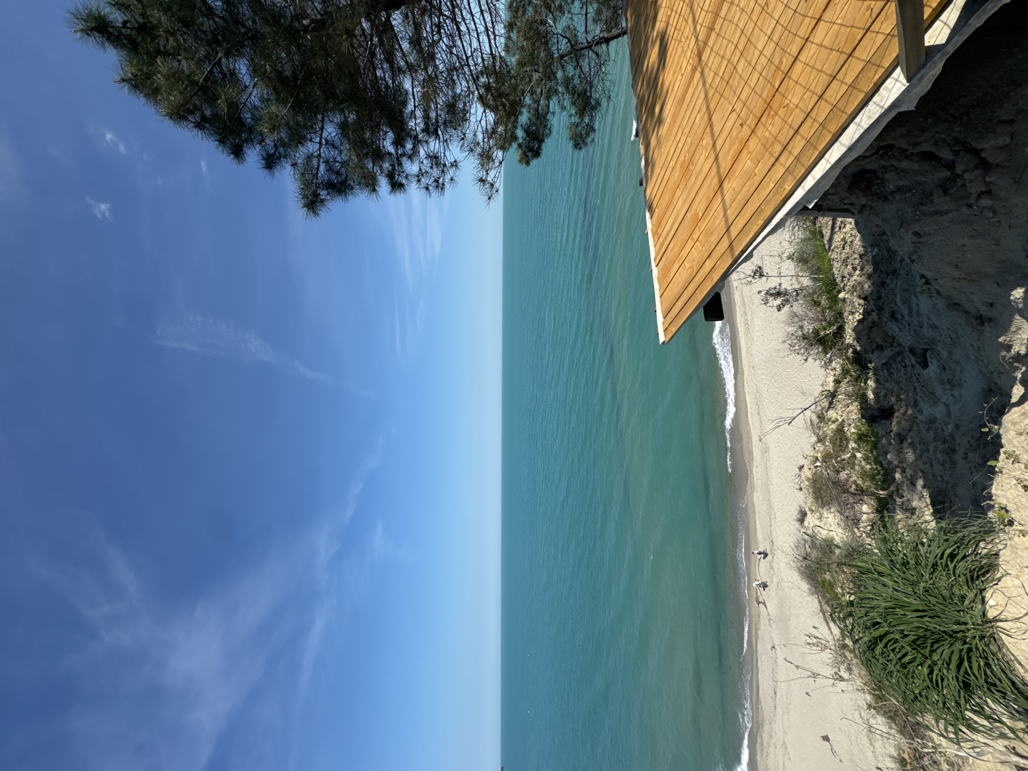 The image shows an elevated view of a beach in Varna, Bulgaria, from a structure on a cliff or a raised platform. The composition is divided into sky, sea, and land.
On the left, the blue sky is partially obscured by tree branches. The sea is a shade of turquoise. A sandy beach is visible along the coastline with small waves.
On the right, a wooden structure, likely part of a building or viewing platform, is seen on the cliffside. The cliff consists of sandy soil and vegetation. There are two people on the beach near the water's edge.