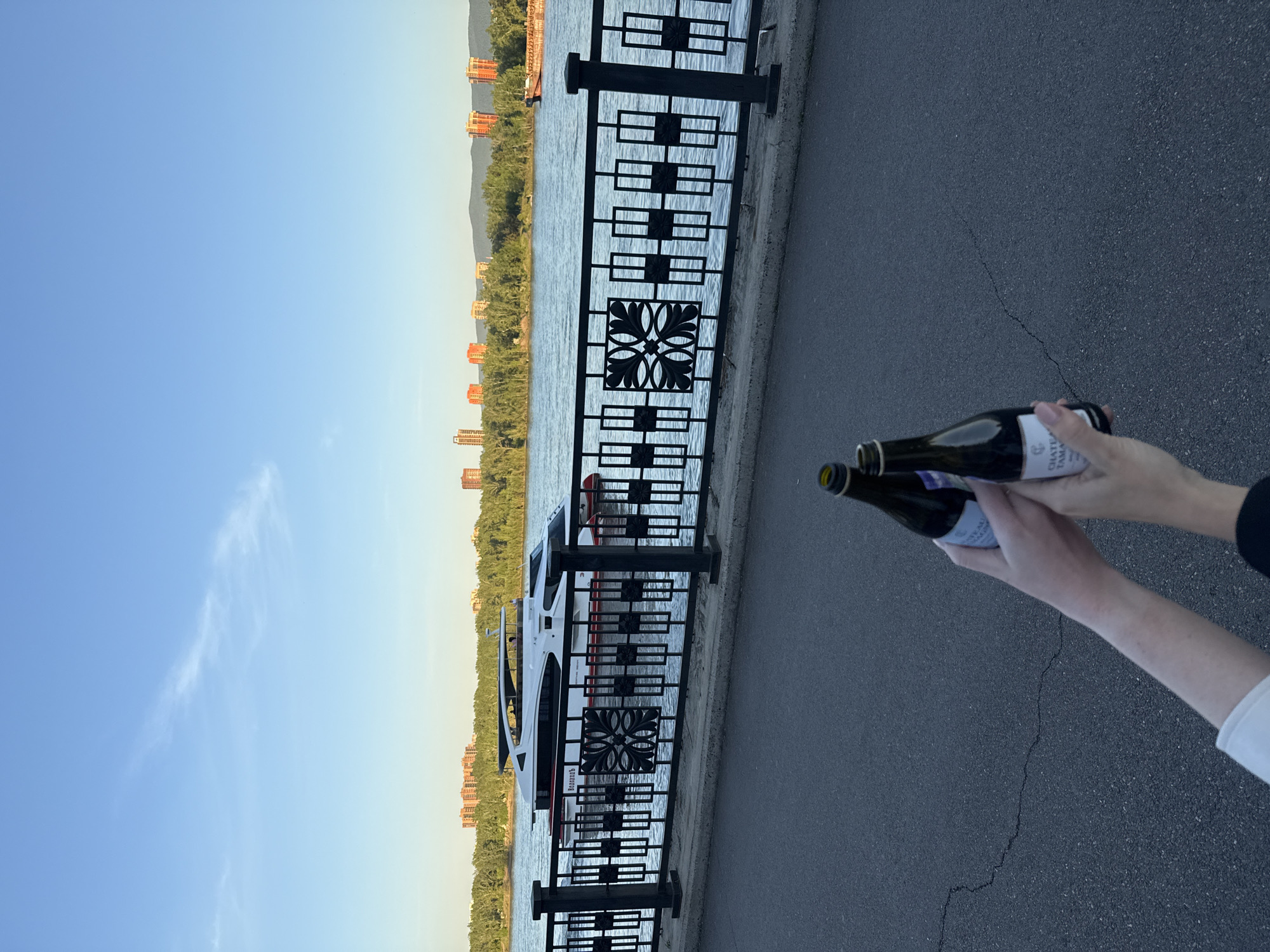 The image, taken in Krasnoyarsk, Russia, shows a scenic view alongside a body of water, likely the Yenisei River, with a focus on two hands holding bottles in the foreground. The perspective is from a person standing on an asphalt path or promenade next to a black metal railing.
In the foreground, two hands are holding two small dark-colored bottles. On the label of the top bottle, the text "CHATEAU TAMA" is visible. The asphalt ground beneath the hands shows a noticeable crack.
Beyond the path and hands, a decorative black metal fence or railing runs horizontally, separating the path from the water. The fence features repeating square patterns with ornamental floral designs. On the water, a large white and red boat or ferry is docked or slowly passing, its side facing the viewer. The name "ВОСХОД" (VOSKHOD), which means "Sunrise" in Russian, is visible on the side of the boat in red lettering.
On the opposite bank of the river, a line of dense green trees and foliage stretches across, with several multi-story buildings, appearing orange/brown in the soft light, interspersed among them and extending towards the distant hills or mountains. The sky is clear and bright blue, suggesting a late afternoon or early evening setting, with faint wisps of white clouds visible in the upper left. The overall scene depicts a calm, pleasant outdoor environment by the river.