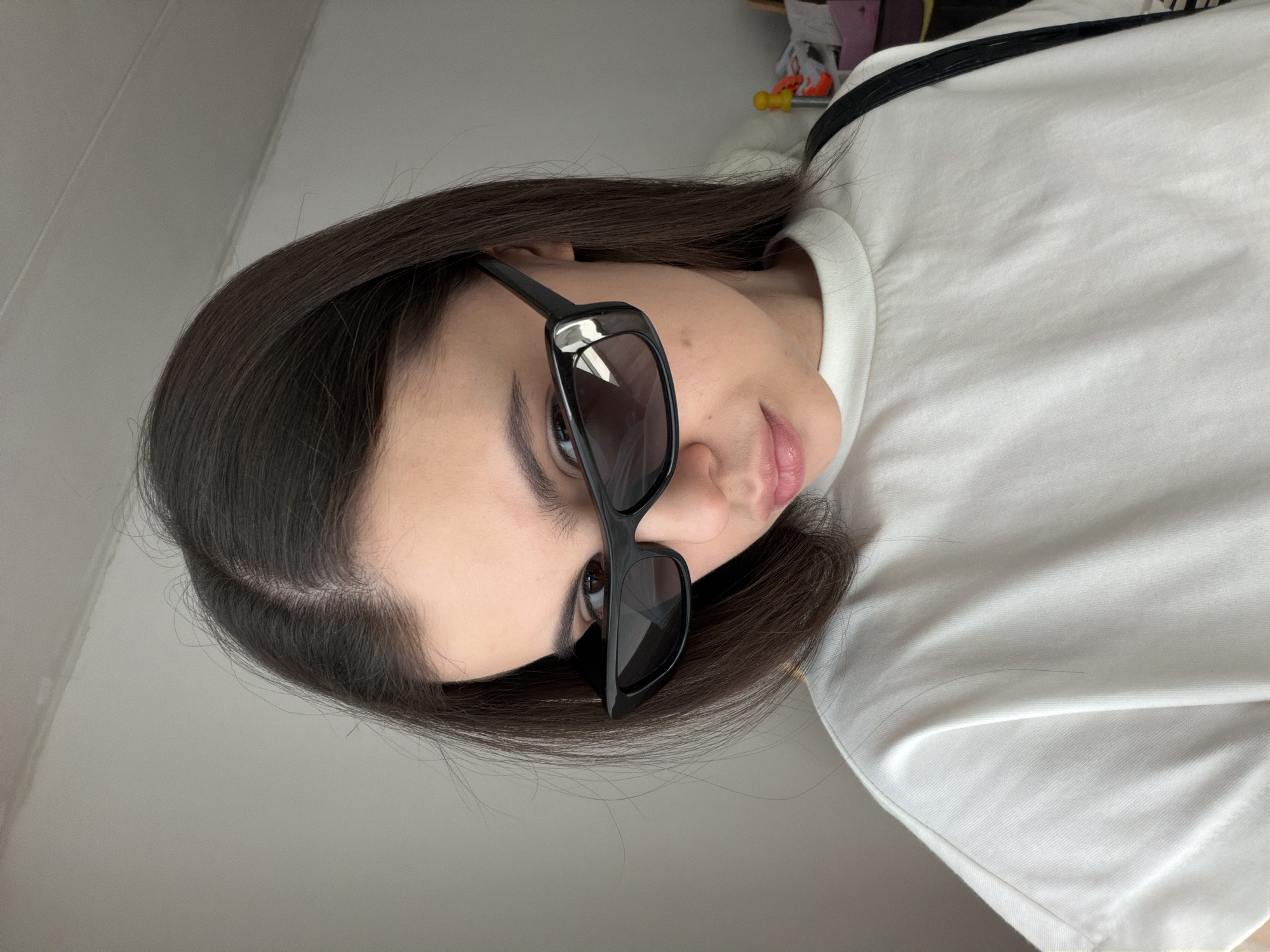 A woman with long, dark hair is wearing black sunglasses and a white t-shirt. The setting appears to be indoors, judging by the wall and objects faintly visible in the background. No specific details suggest the location is Chaikovsky, Russia.