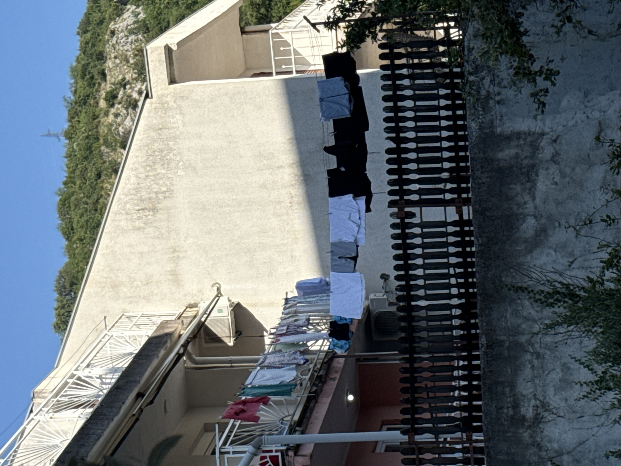 In Rafailovići, Montenegro, laundry is hung on a clothesline suspended between two buildings. The clothesline is laden with an assortment of garments, including shirts, pants, and other items of clothing. The buildings themselves appear to be multi-story structures with balconies and windows. A wooden fence runs horizontally across the lower portion of the image, separating the foreground from the buildings. In the background, a lush green hillside rises, dotted with rocks and trees, under a clear blue sky. A power pylon is visible on the top of the hill.